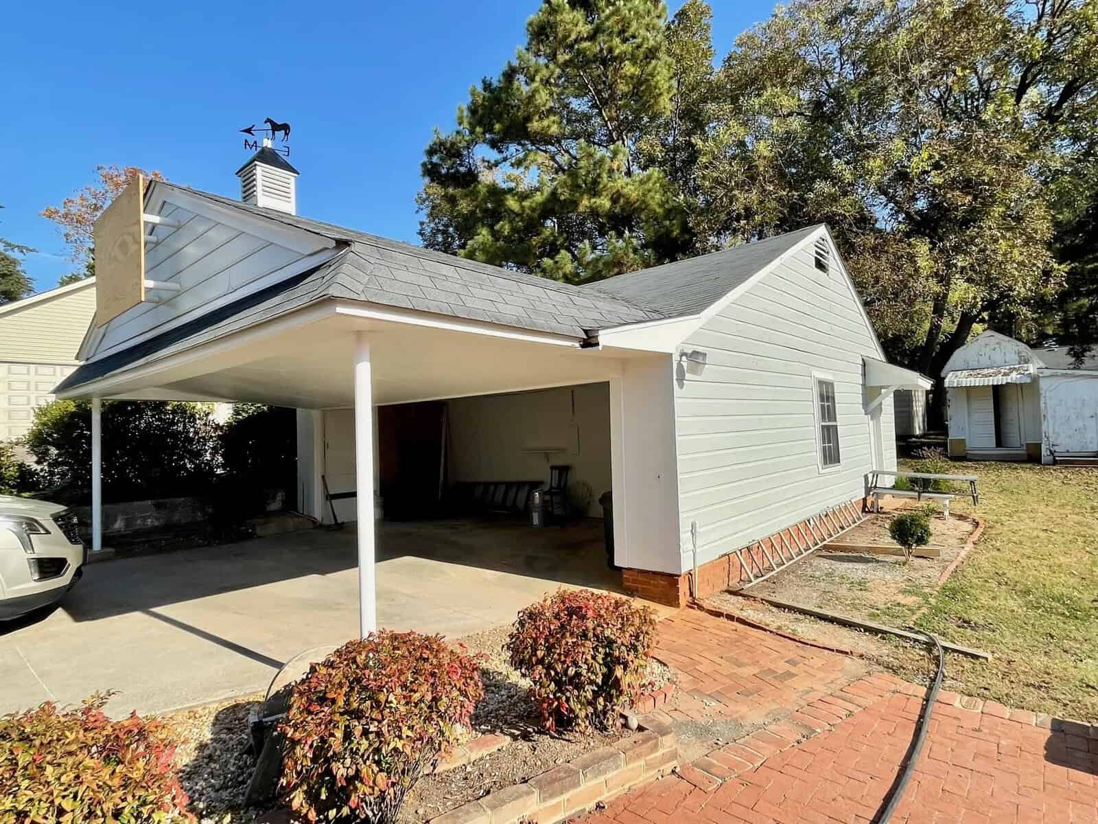 A single-story house with white siding featuring a carport on the left, a brick pathway leading to the front entrance, and a small garden area with shrubs in the foreground.