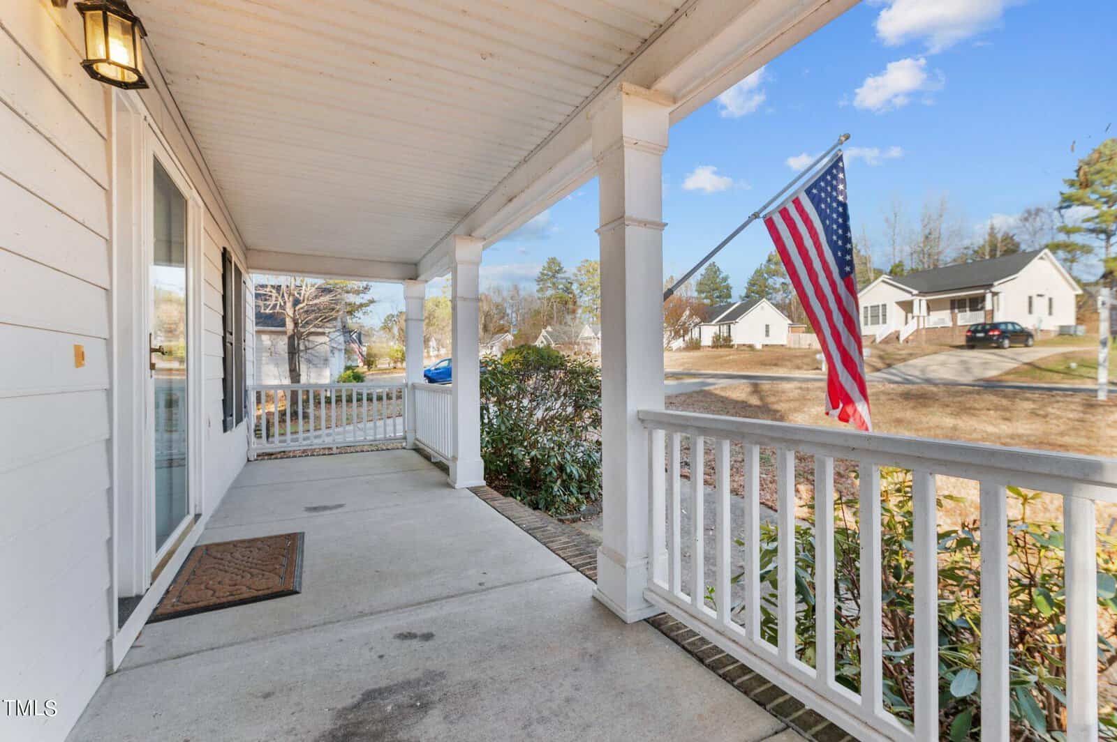 A suburban front porch with white railings and a welcome mat. An American flag is displayed on a post. The background shows a quiet street with houses and trees under a clear blue sky.