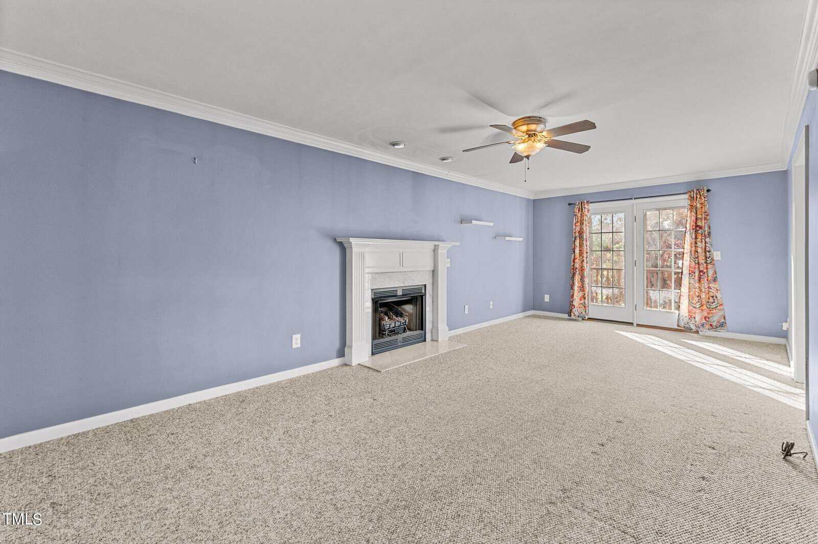 A spacious living room with light blue walls features a white fireplace, beige carpet, a ceiling fan with lights, and French doors leading to an outdoor area with floral curtains. Shelving units are installed on the wall near the fireplace.