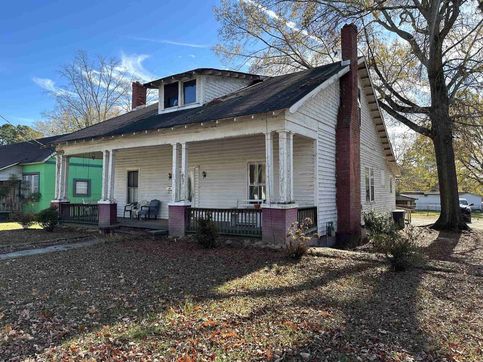 An old, weathered single-story house with a porch and missing roof sections at 312 E Andrews Avenue, Henderson NC, stands neglected, surrounded by fallen leaves and bare trees under a clear blue sky