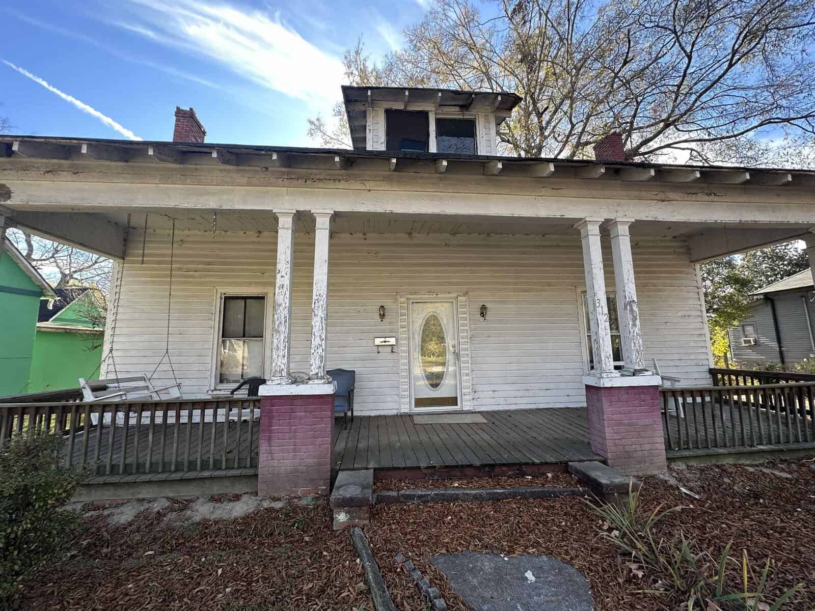An older single-story house showing signs of wear, with a large front porch supported by columns and a peeling paint exterior.