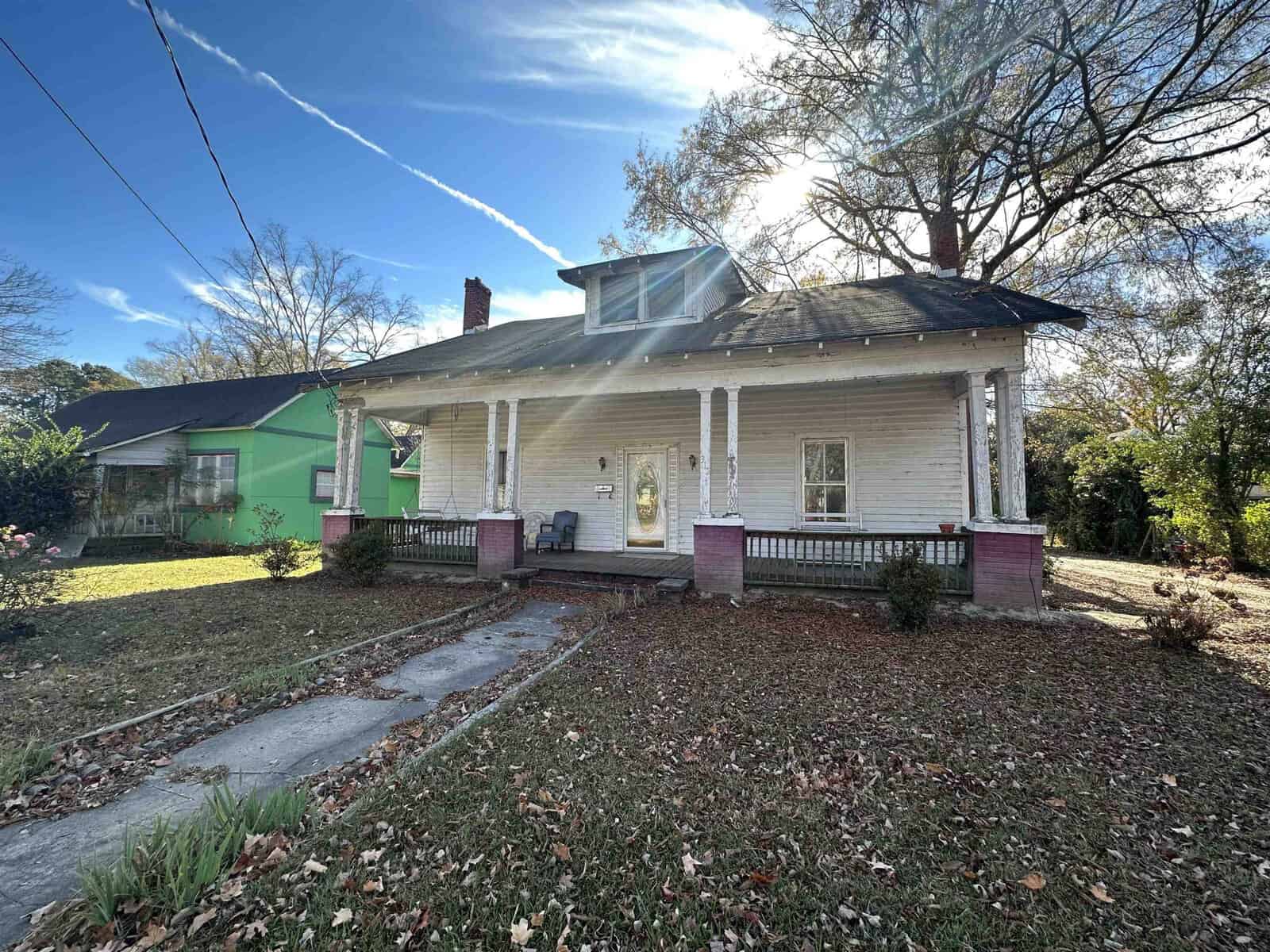A single-story house with white siding, a front porch with pink accents, and a sloping front yard under a clear sky with sun glare visible.