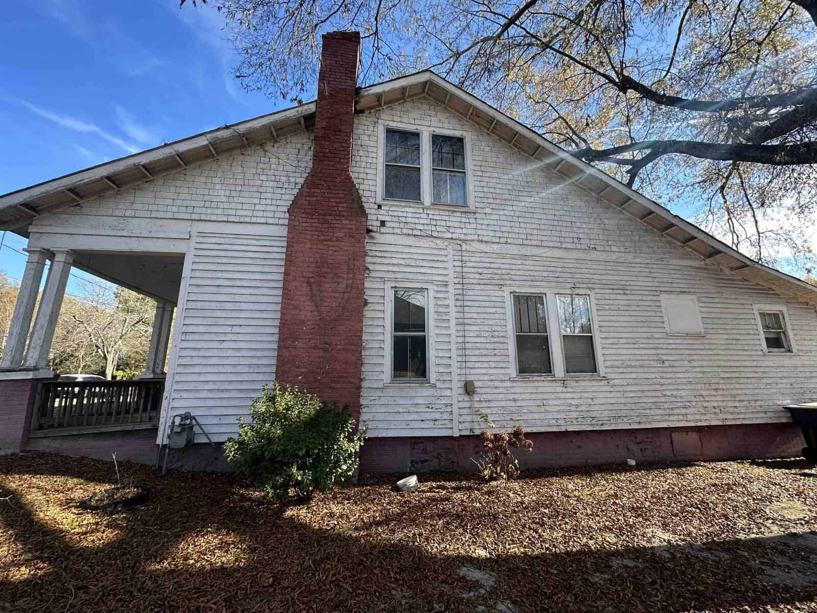 An old white house with peeling paint, a red chimney, and a covered porch, surrounded by leafless trees and a blanket of fallen leaves.