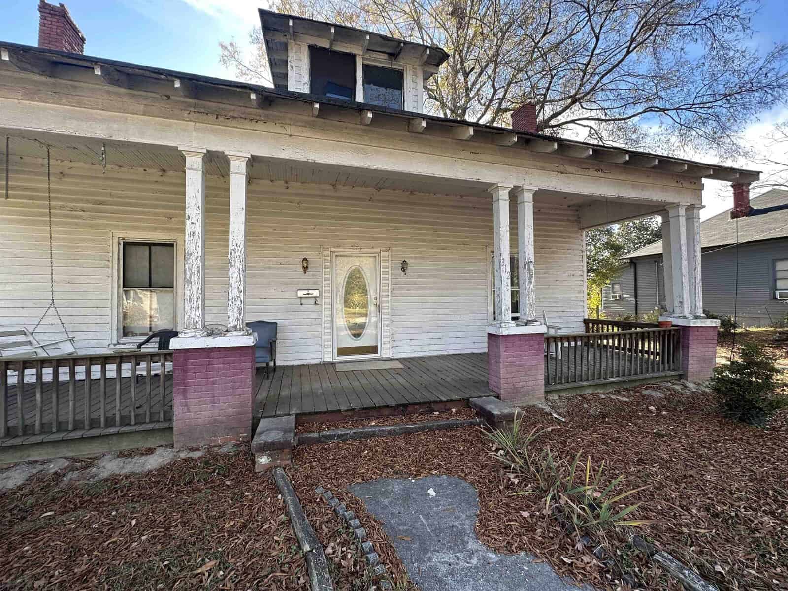 White wooden house with a covered porch, featuring decorative columns and a windowed front door, surrounded by a yard with leaf-littered grass and a concrete pathway.