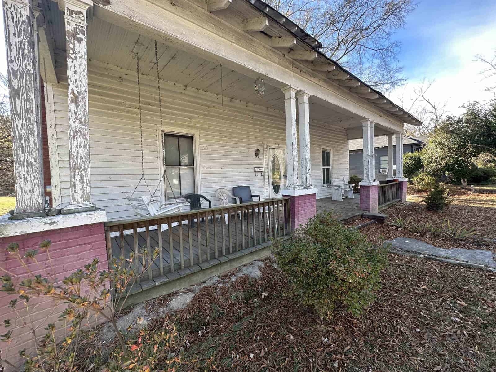 An old, weathered house with a front porch showing signs of neglect and disrepair, featuring peeling paint, overgrown bushes, and a couple of chairs.