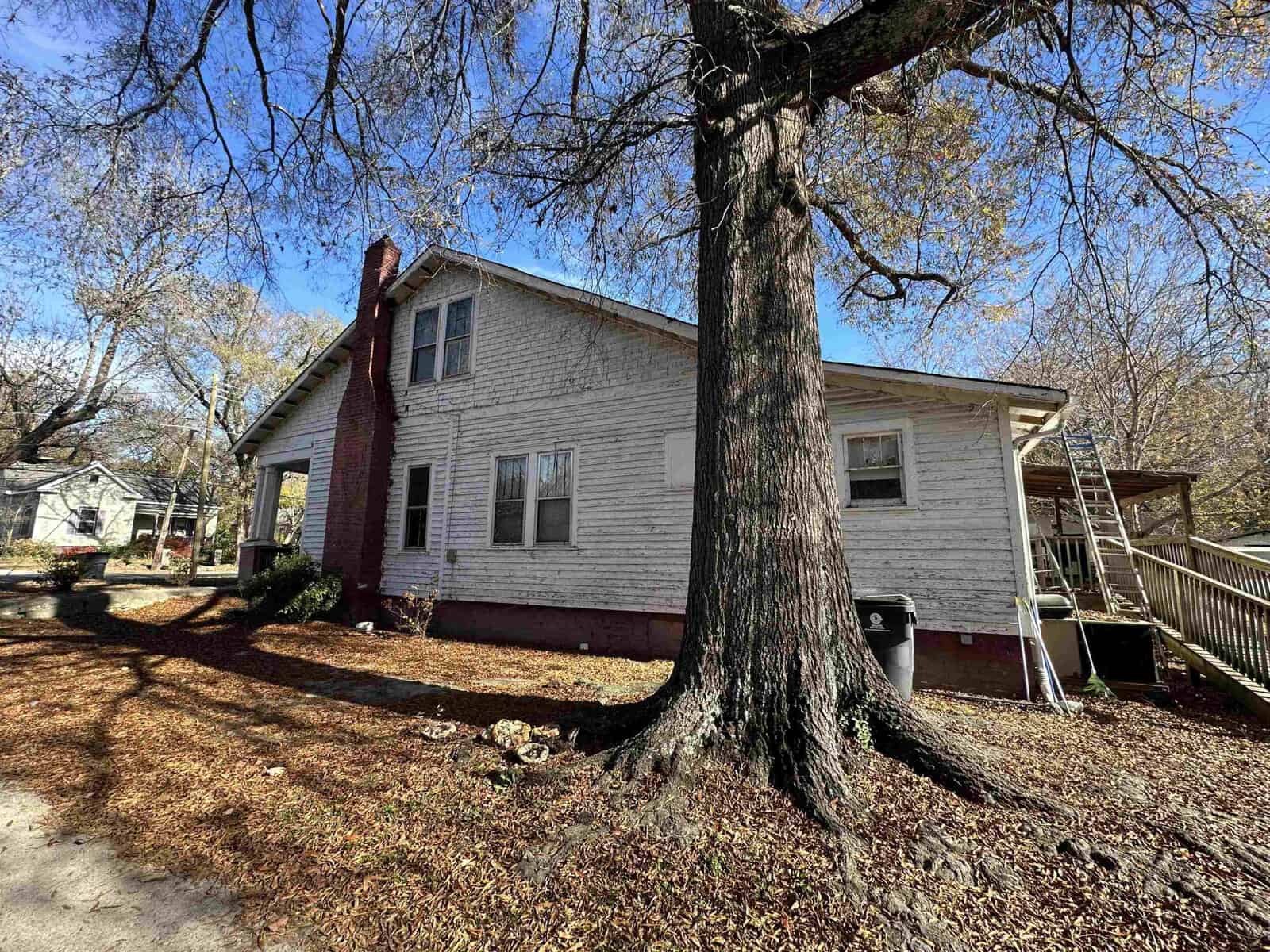 A two-story white house with red trim and a large tree in the front yard on a sunny day with shadows from the bare branches on the ground.