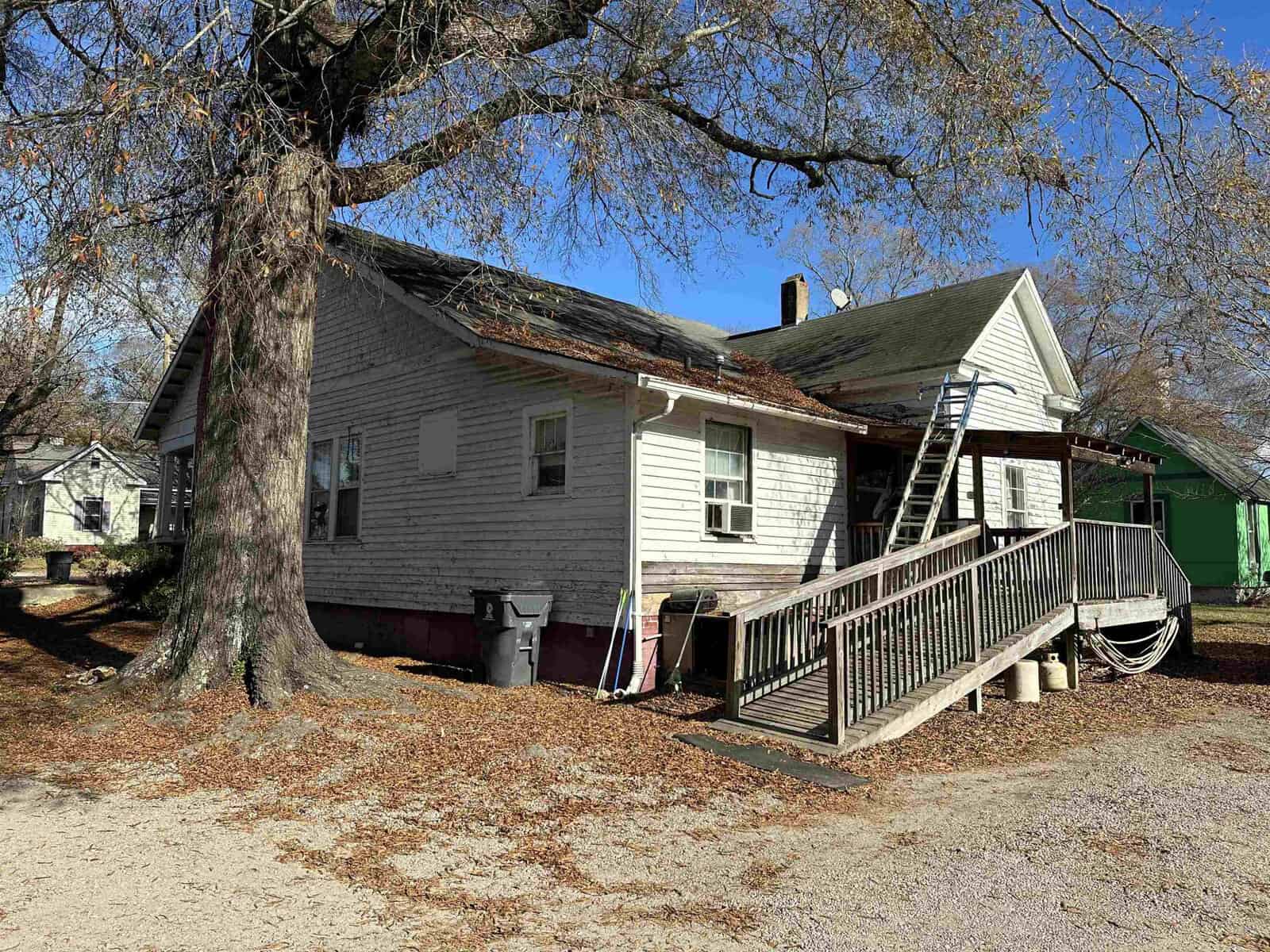 A modest, weathered single-story house with a wooden porch and a tree shedding leaves in the yard.
