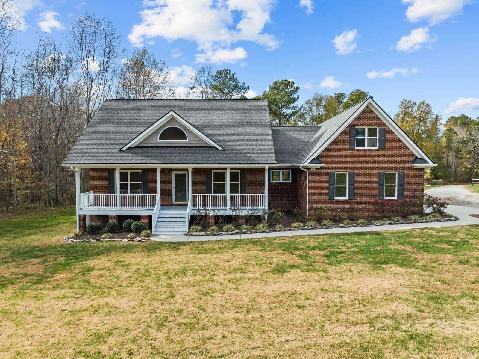 The red brick house at 1670 Piney Creek Lane boasts a gray roof and sits in a grassy area. It features a front porch with white railings and steps, surrounded by shrubs and trees under a partly cloudy blue sky, enhancing its warm, welcoming charm.