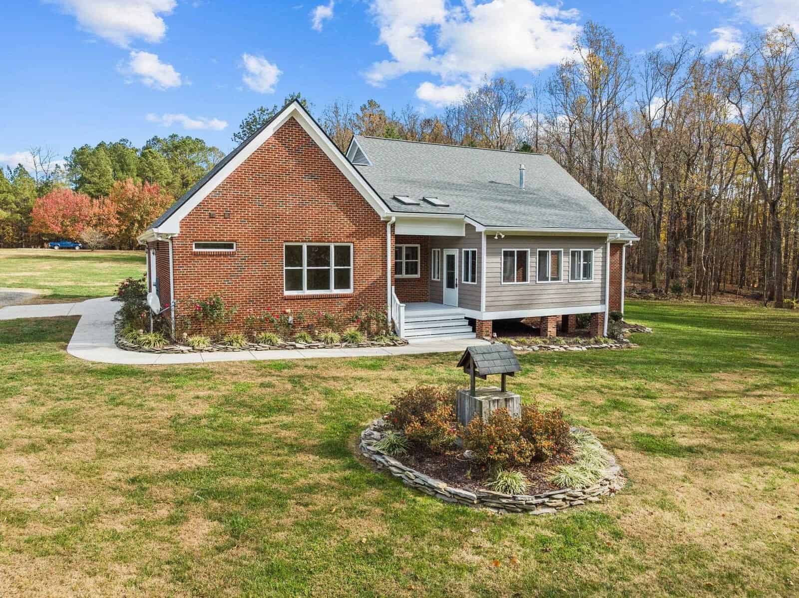 A brick house with a gray roof sits in a grassy yard surrounded by trees under a blue sky. A small garden with shrubs and a wooden well is in the foreground. The house features several windows and a driveway leading to the entrance.