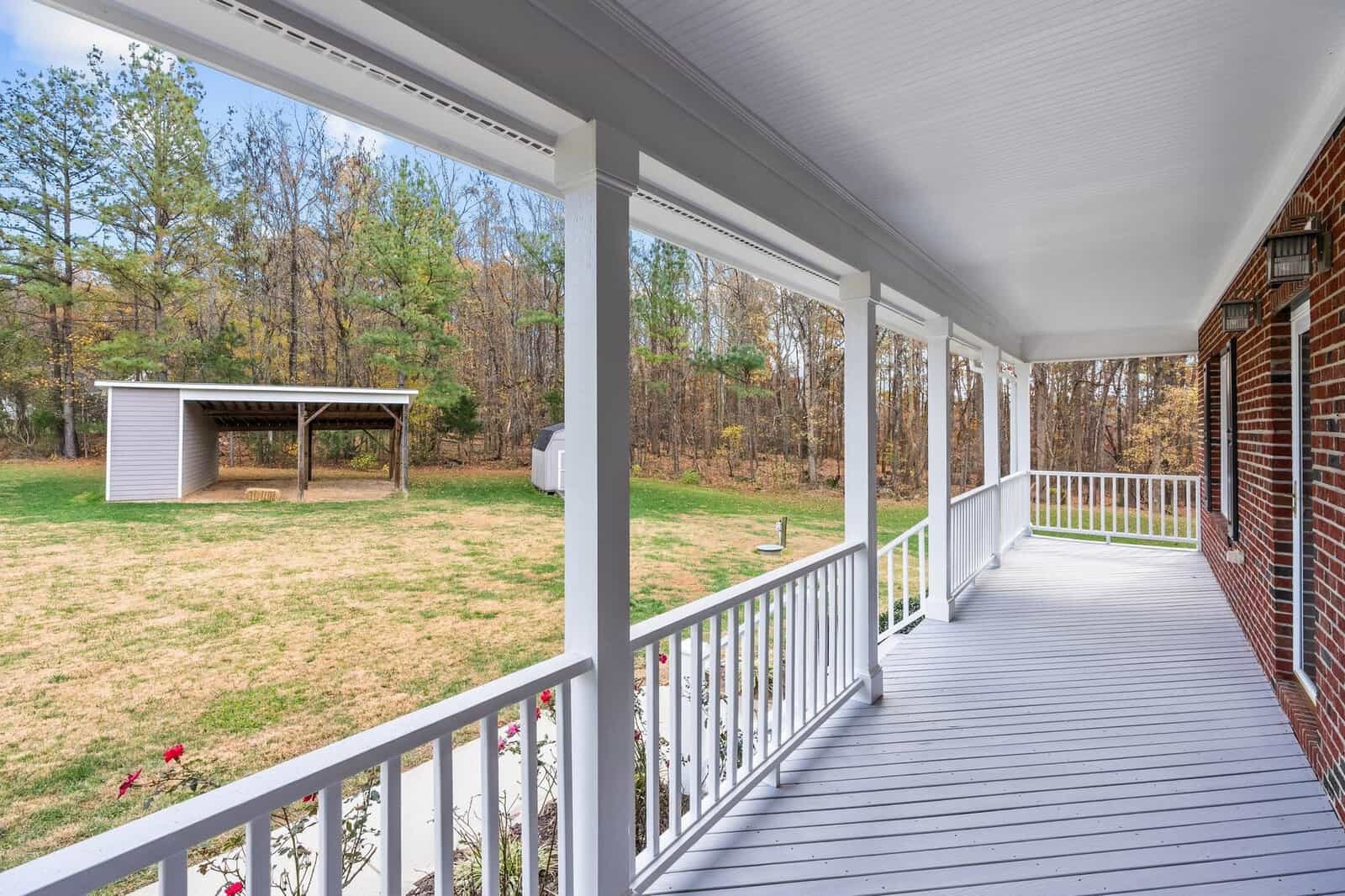 A spacious white porch with wooden railings overlooks a grassy backyard with a small shed and carport. Tall trees surround the area, and blooming rose bushes line the porch. The sky is clear and sunny.