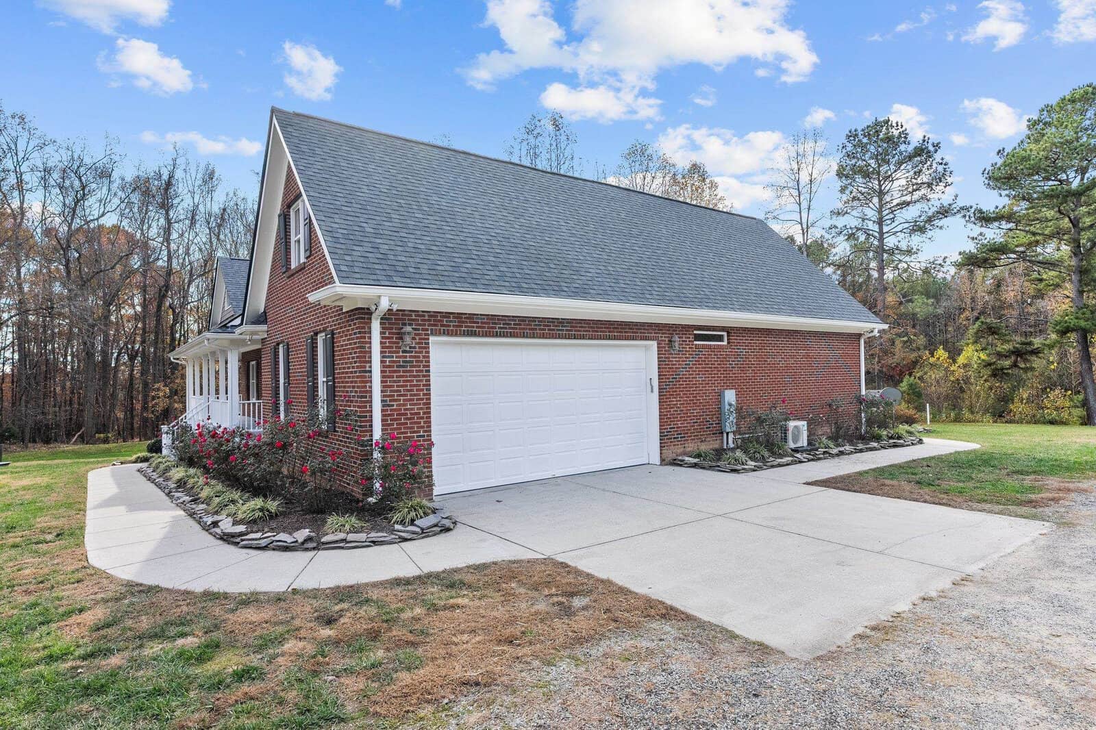 A red brick house with a dark gray roof and a white garage door. It is surrounded by a landscaped garden with flowers and shrubs. The driveway is paved, and there's a backdrop of trees under a partly cloudy blue sky.