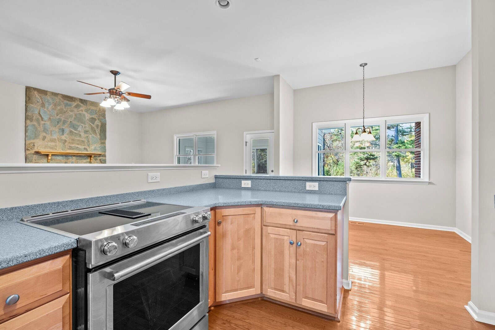 A bright kitchen features wooden cabinets, a stainless steel stove, and a blue-gray countertop. An open space reveals a living area with a stone fireplace and ceiling fan, while a window offers a view of greenery outside.