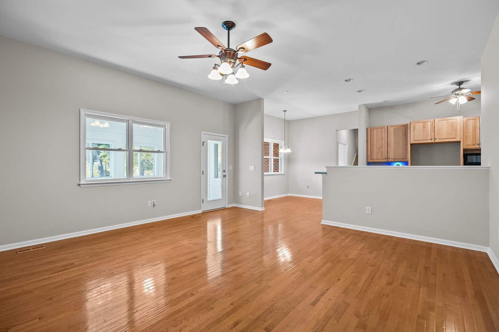 Spacious, empty living area with polished hardwood floors, light gray walls, and a ceiling fan with lights. Large windows let in natural light. In the background, there's a kitchen with wooden cabinets and a breakfast bar.