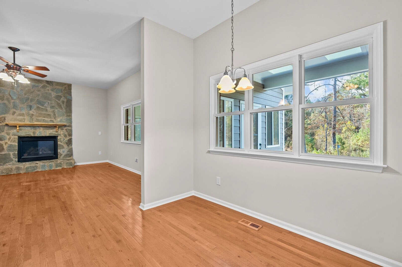 A bright, empty room with wooden floors and large windows, overlooking a scenic outdoor area. A stone fireplace is on the left, and a ceiling fan hangs above. A three-light pendant fixture is near the windows.