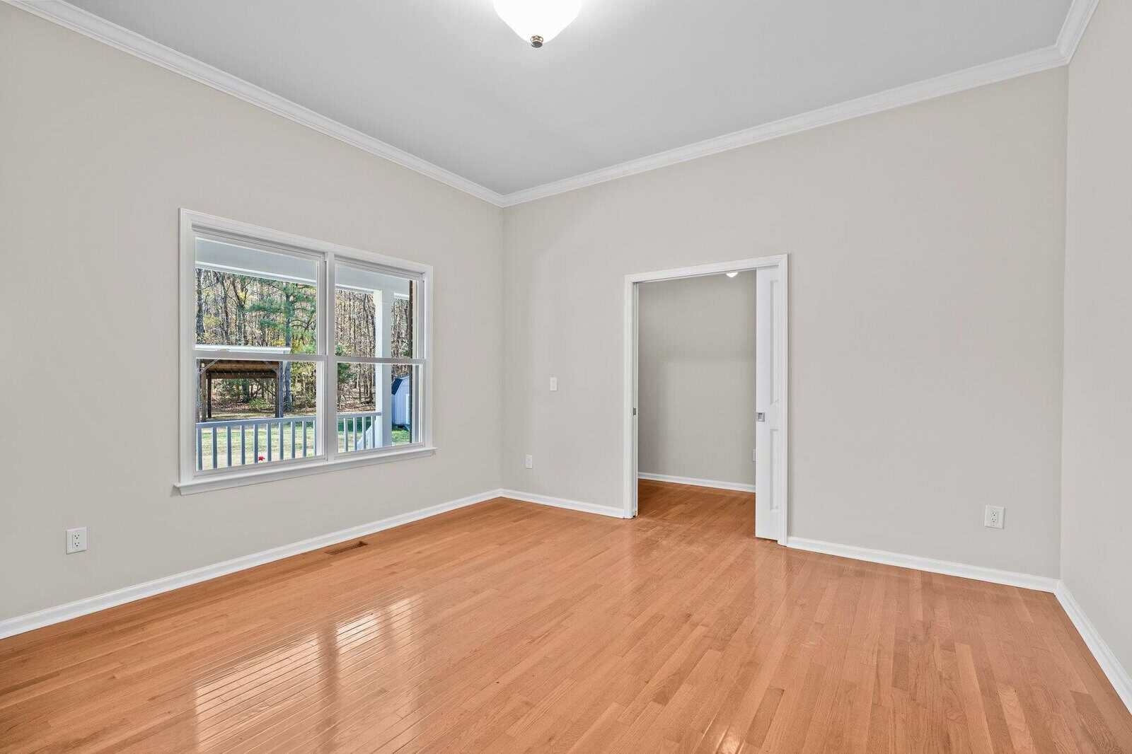 An empty room with light wood flooring, beige walls, and a white ceiling. A window on the left lets in natural light and offers a view of trees. The room has a doorway leading to another space.
