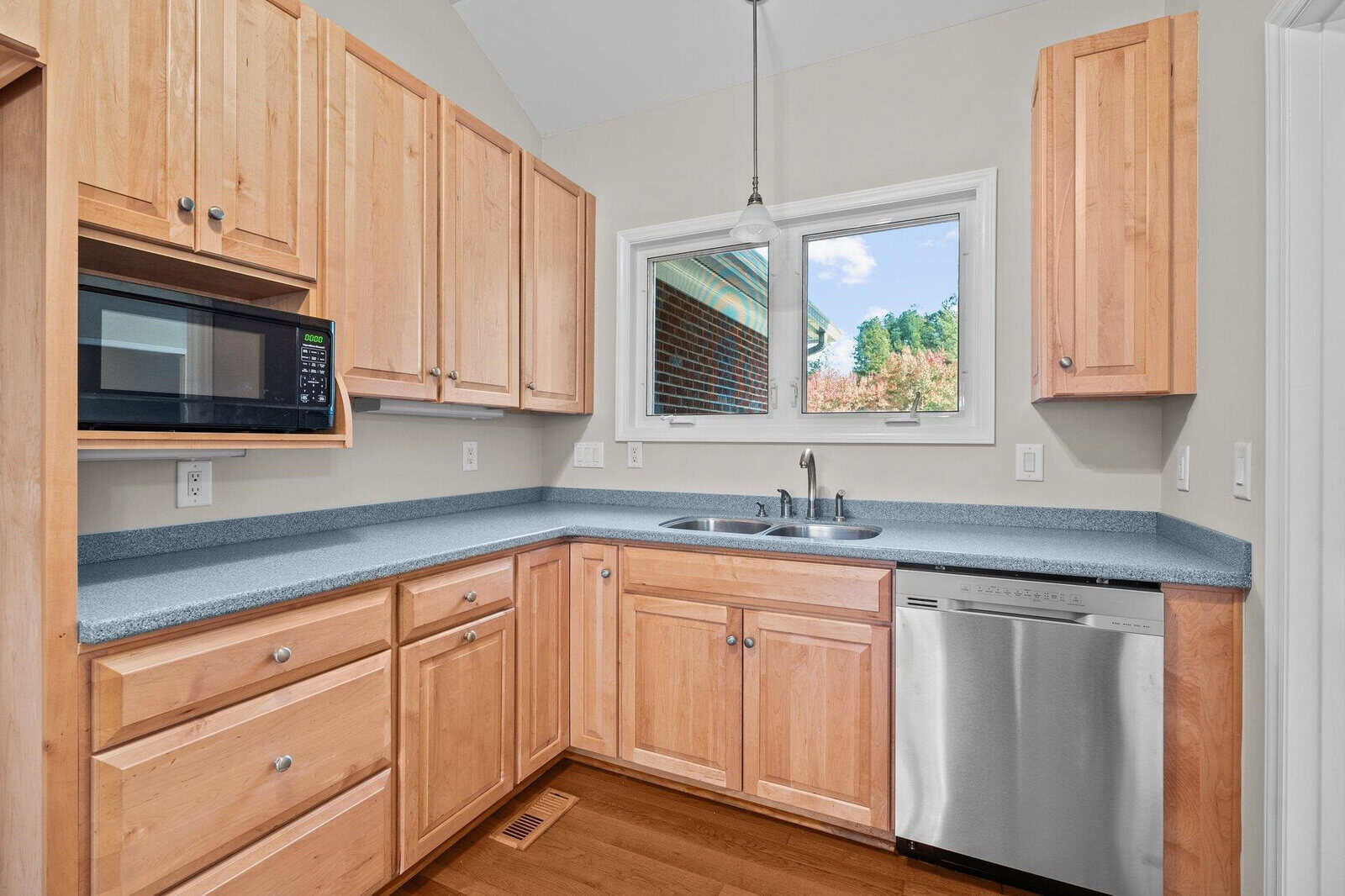 A kitchen with wooden cabinets, a gray countertop, and stainless steel dishwasher. A microwave sits above the counter. A single window provides natural light. The walls are light-colored, and the floor is wooden.