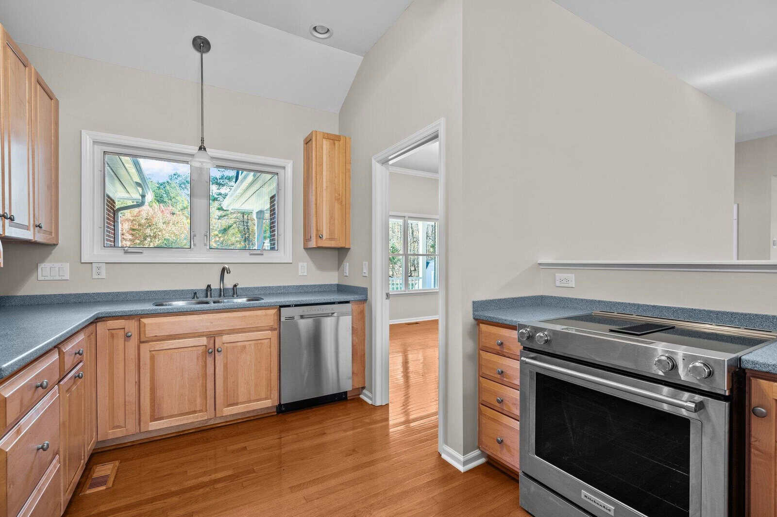 A modern kitchen with light wood cabinets, stainless steel dishwasher, and an electric stove. There's a window over the sink and hardwood flooring. A doorway opens to another room, visible in the background.