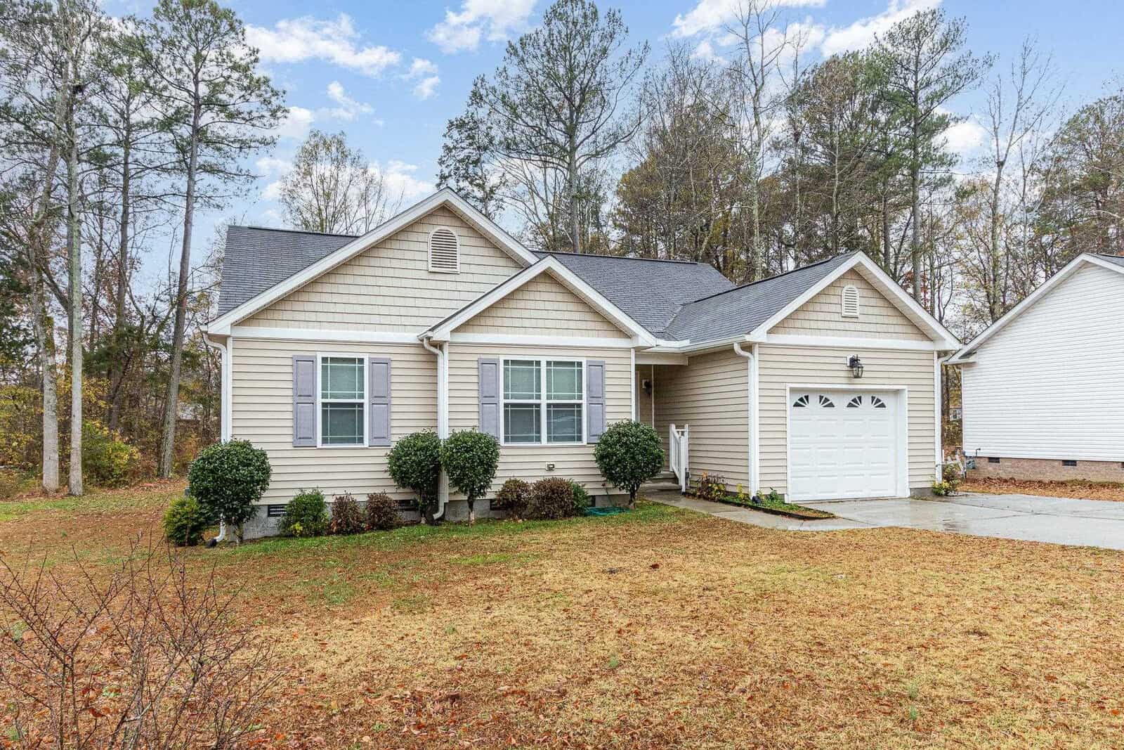 A single-story beige house with a gray shingled roof, a white garage door, and a small front porch. The house is set against a backdrop of tall trees, and the front yard is grassy with some shrubs near the entrance. The sky is cloudy.