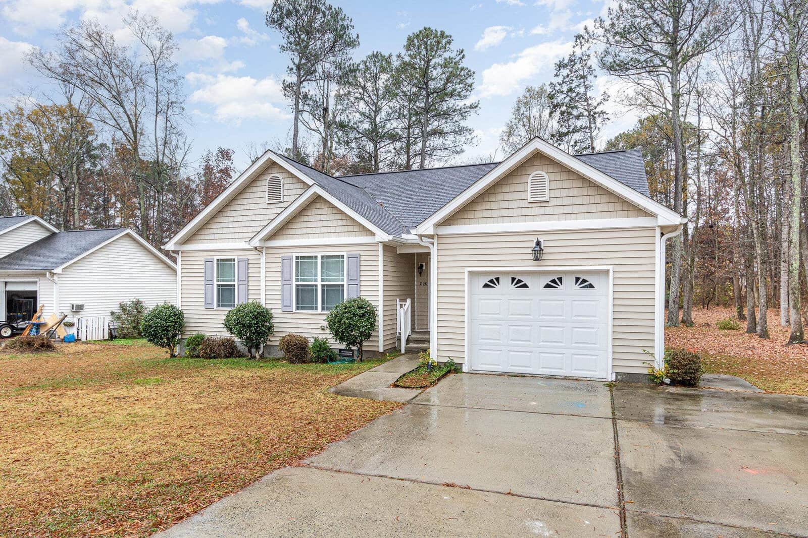 Single-story beige house with white trim and a gable roof. It has a one-car garage, three front windows, and is surrounded by trees. The driveway and grass lawn are wet, indicating recent rain. Cloudy sky in the background.