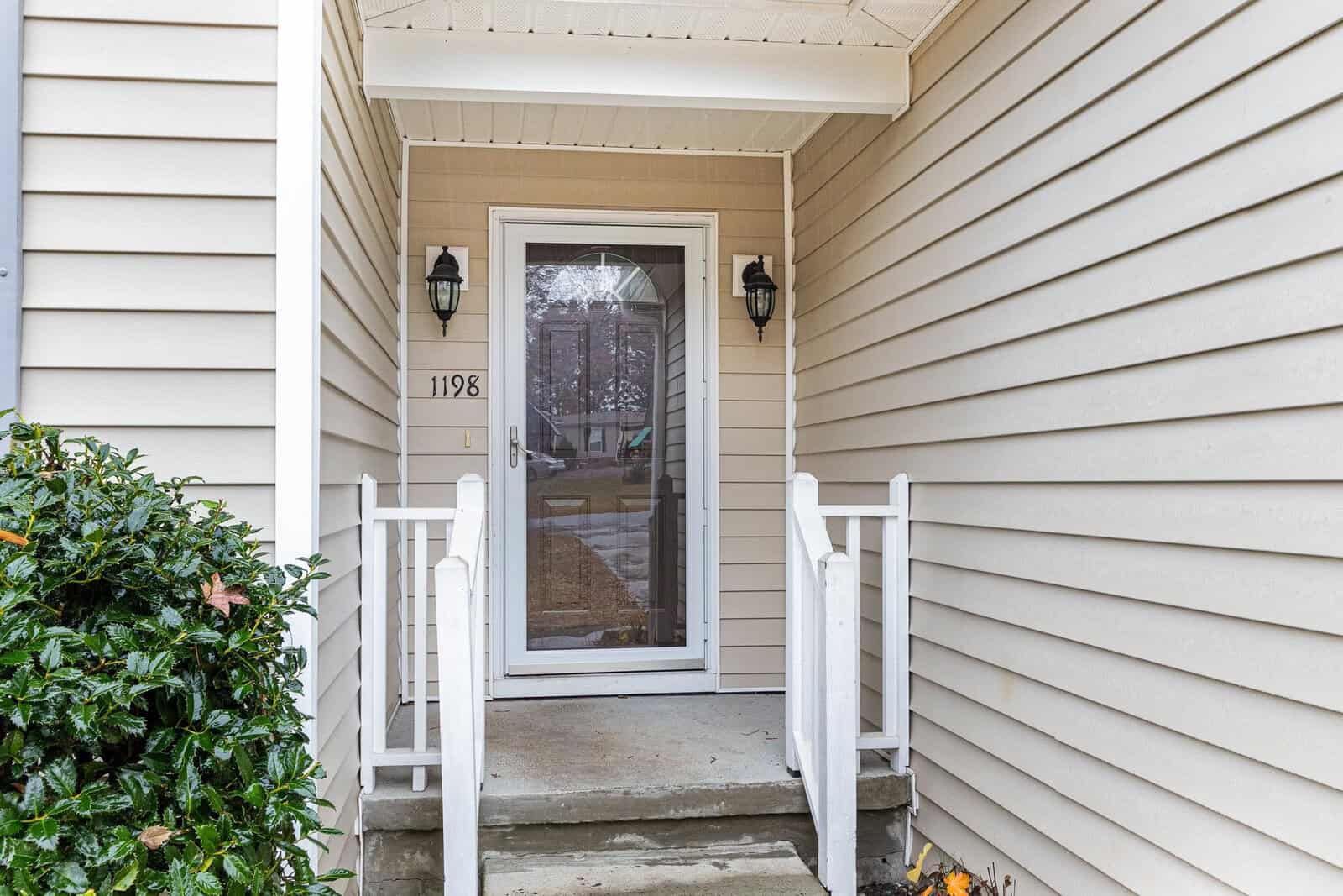 A front entryway of a house with beige siding, featuring a glass storm door, two black wall lanterns, and white railings on both sides of a small concrete porch. There are green bushes on the left side.