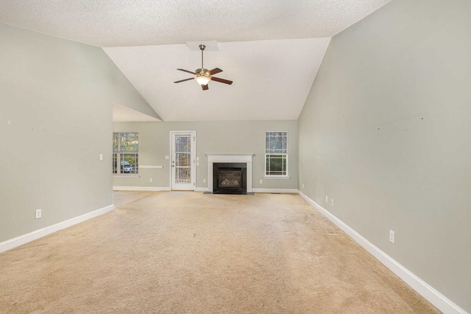 A spacious living room with beige carpet, light gray walls, and a high ceiling. It features a central ceiling fan and a white-framed fireplace between two tall windows. The room leads to an adjacent area with large windows.