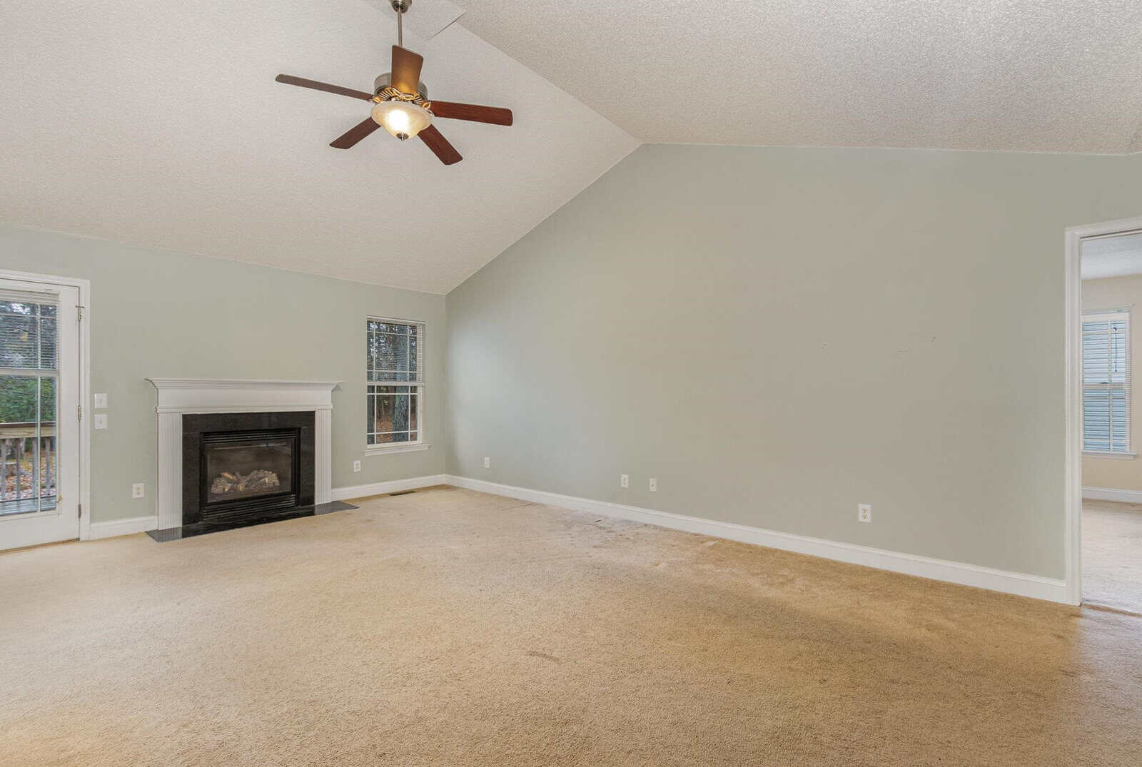 A spacious living room with beige carpet, light green walls, and a high ceiling. The room features a ceiling fan, a fireplace, a glass door leading outside, and a large window. An open doorway connects to another room on the right.