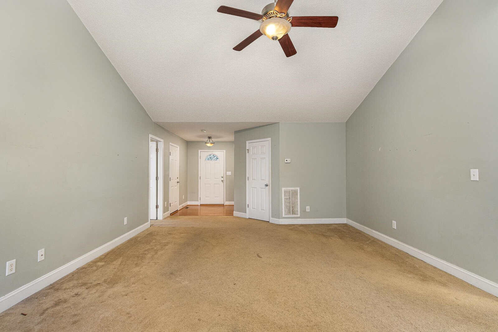 A spacious empty room with light green walls, a beige carpet, and a ceiling fan with wooden blades. There are multiple white doors, including a front door with a small window. The ceiling is vaulted, adding to the open feel of the space.