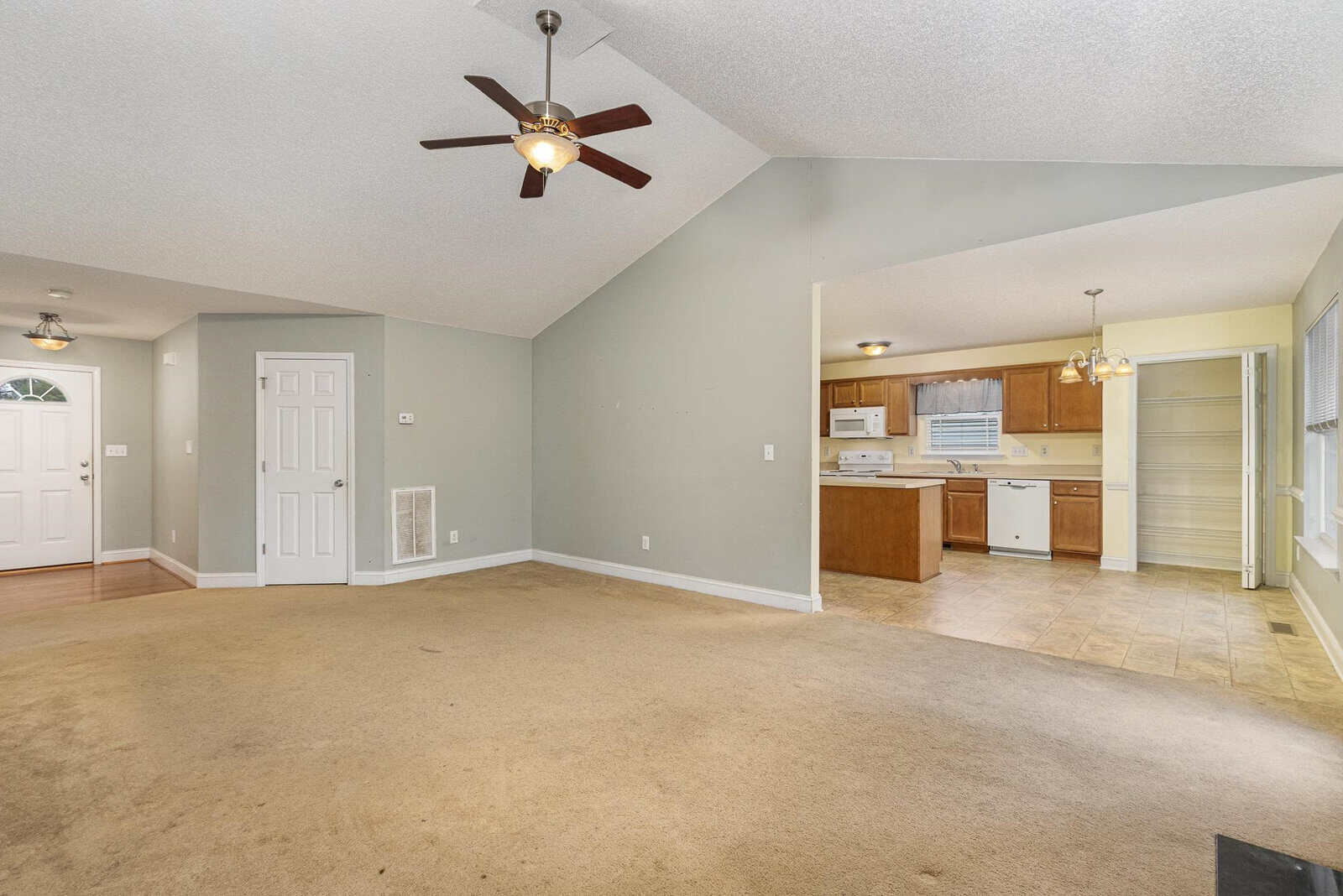 Spacious living area with beige carpet, ceiling fan, and light green walls. Open transition to a kitchen with wooden cabinets, white appliances, and tiled floor. Adjacent dining area with large windows offering natural light.