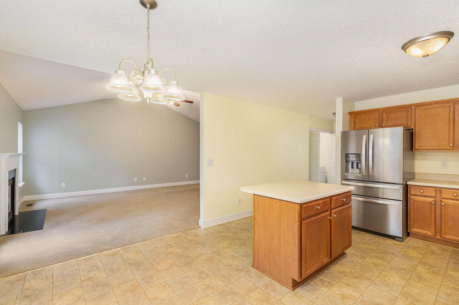 A kitchen with an island and wooden cabinets, featuring stainless steel appliances. An open living area with a fireplace is visible in the background. The flooring is tile in the kitchen and carpet in the living area. A hanging light fixture is present.