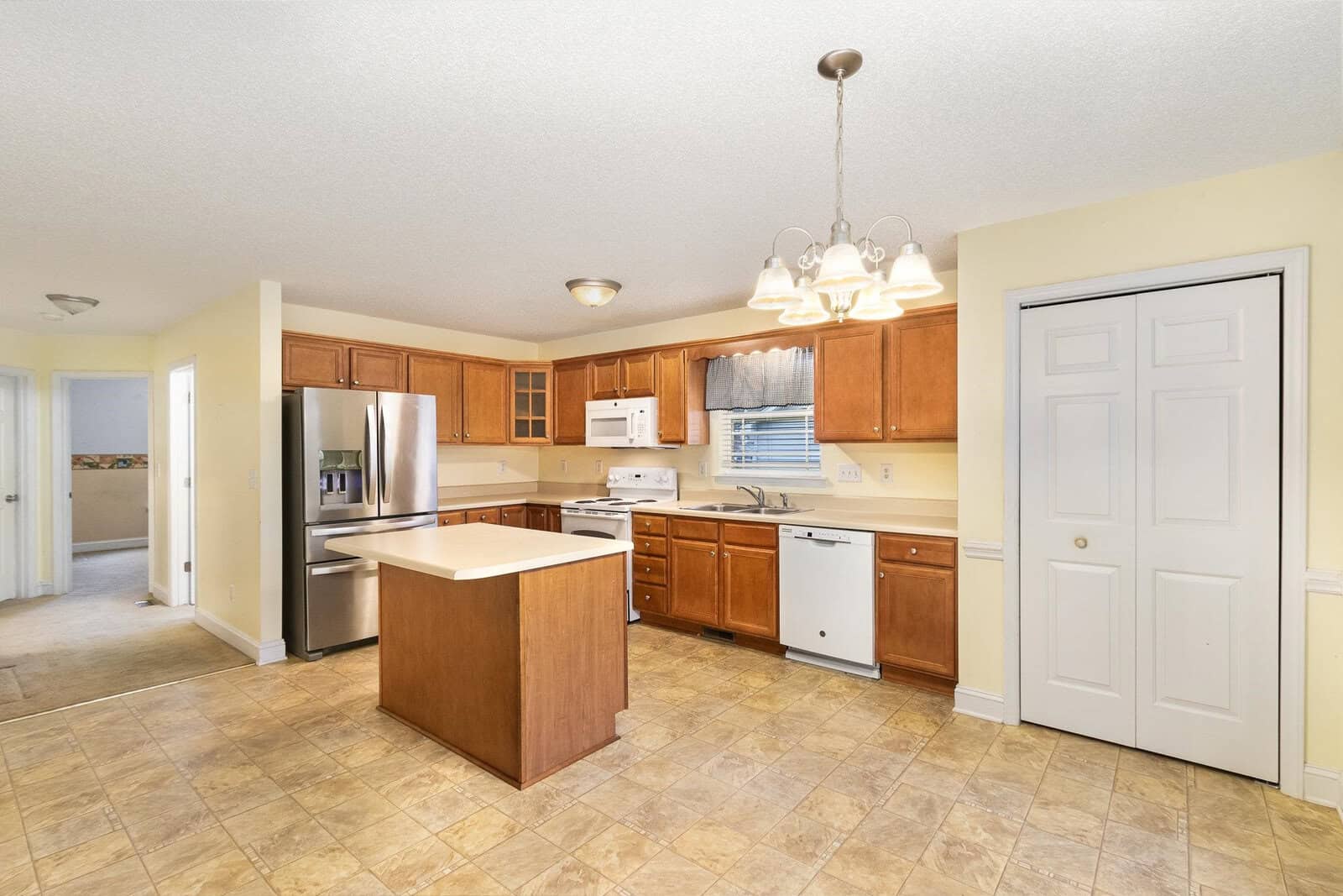 A spacious kitchen with wooden cabinets, a central island, and stainless steel appliances. The floor is tiled, and there's a door on the right. A light fixture hangs from the ceiling, and soft yellow walls provide a warm ambiance.