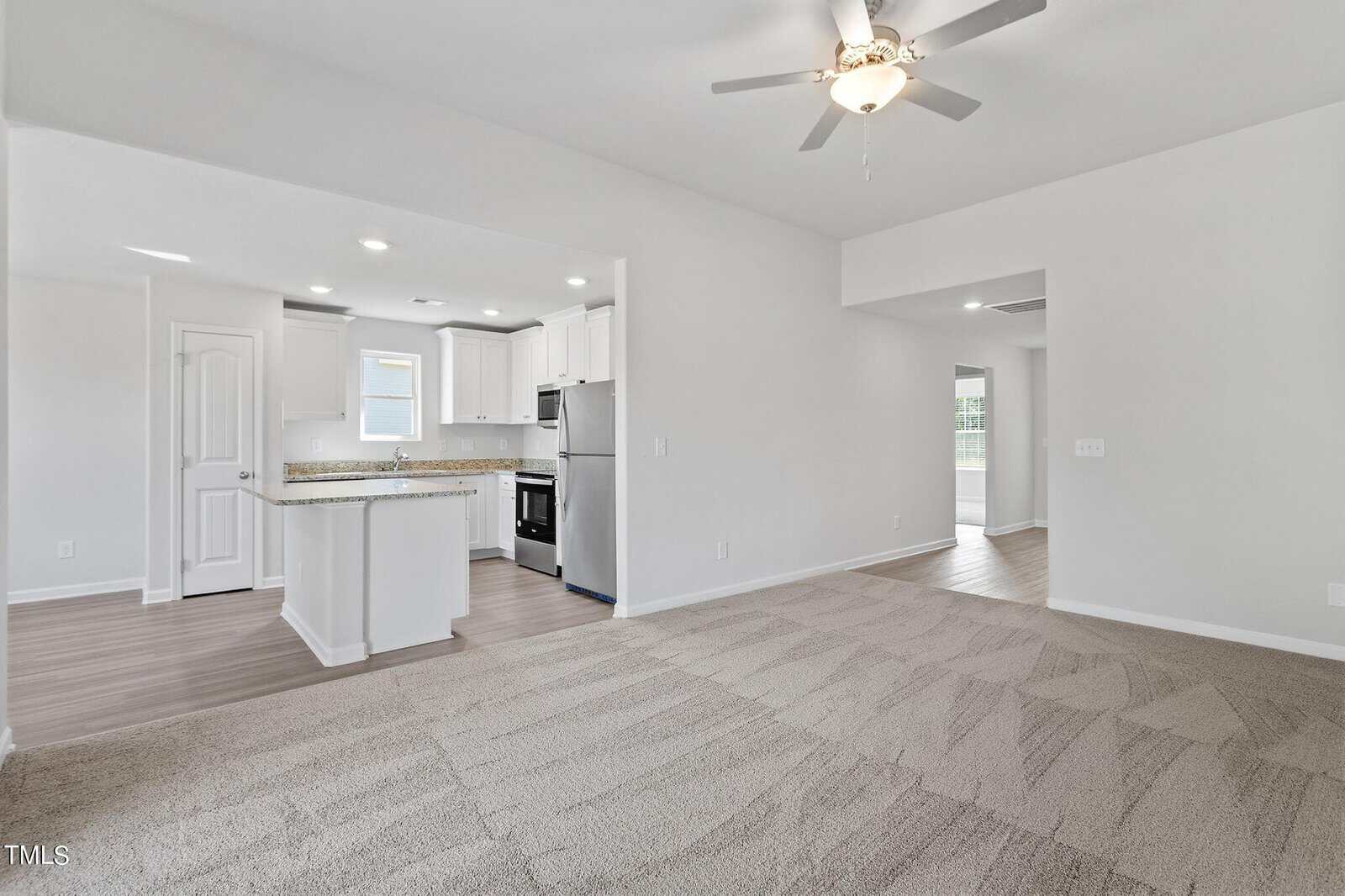 Spacious, unfurnished room with beige carpet and white walls. The open floor plan includes a kitchen with white cabinets, stainless steel appliances, and hardwood floors. A ceiling fan hangs above the living area. Natural light enters through windows.