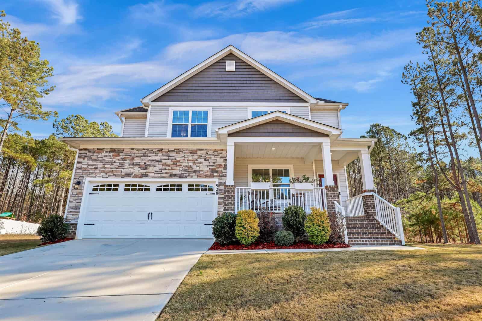 Nestled on 920 Weatherby Lane, this two-story Creedmoor home features a brick and stone facade, a two-car garage, and a welcoming porch with white railings. It's surrounded by landscaped bushes and a green lawn, with tall trees set against the backdrop of a clear blue sky.