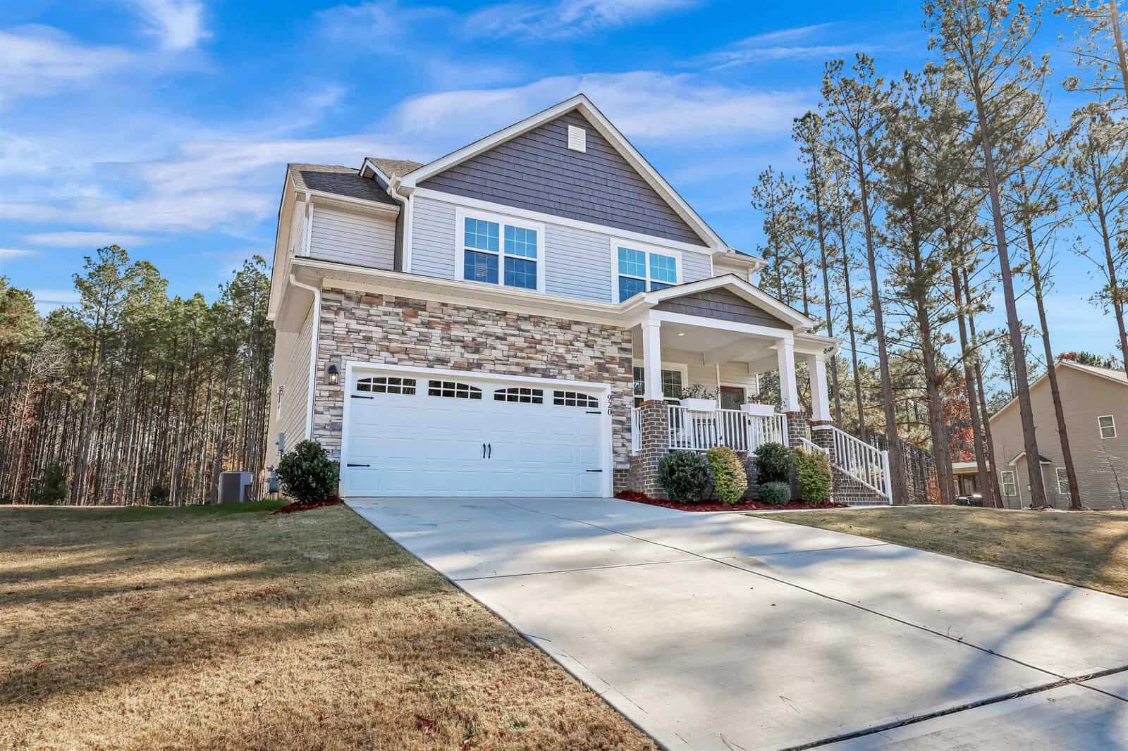 A two-story house with a stone and siding facade is shown. It features a two-car garage, a covered front porch, and sits on a sloped driveway. The house is surrounded by neatly manicured bushes and trees under a clear blue sky.