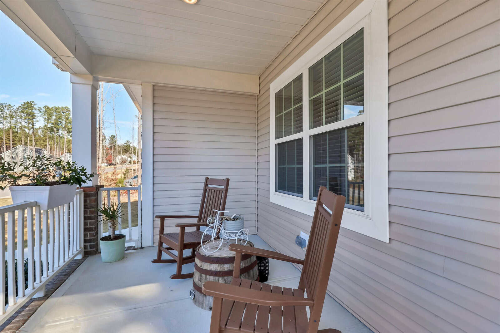 A cozy porch with two wooden rocking chairs and a small round table featuring a decorative bicycle. The porch overlooks a yard with trees, and there is a white railing and a potted plant in the corner. The siding of the house is beige.