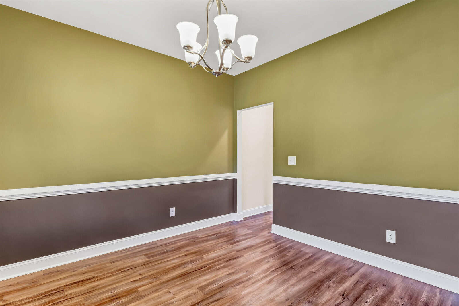 An empty room with olive green and brown walls, white trim, and wooden flooring. A white, modern chandelier hangs from the ceiling. There is a doorway leading to another area.