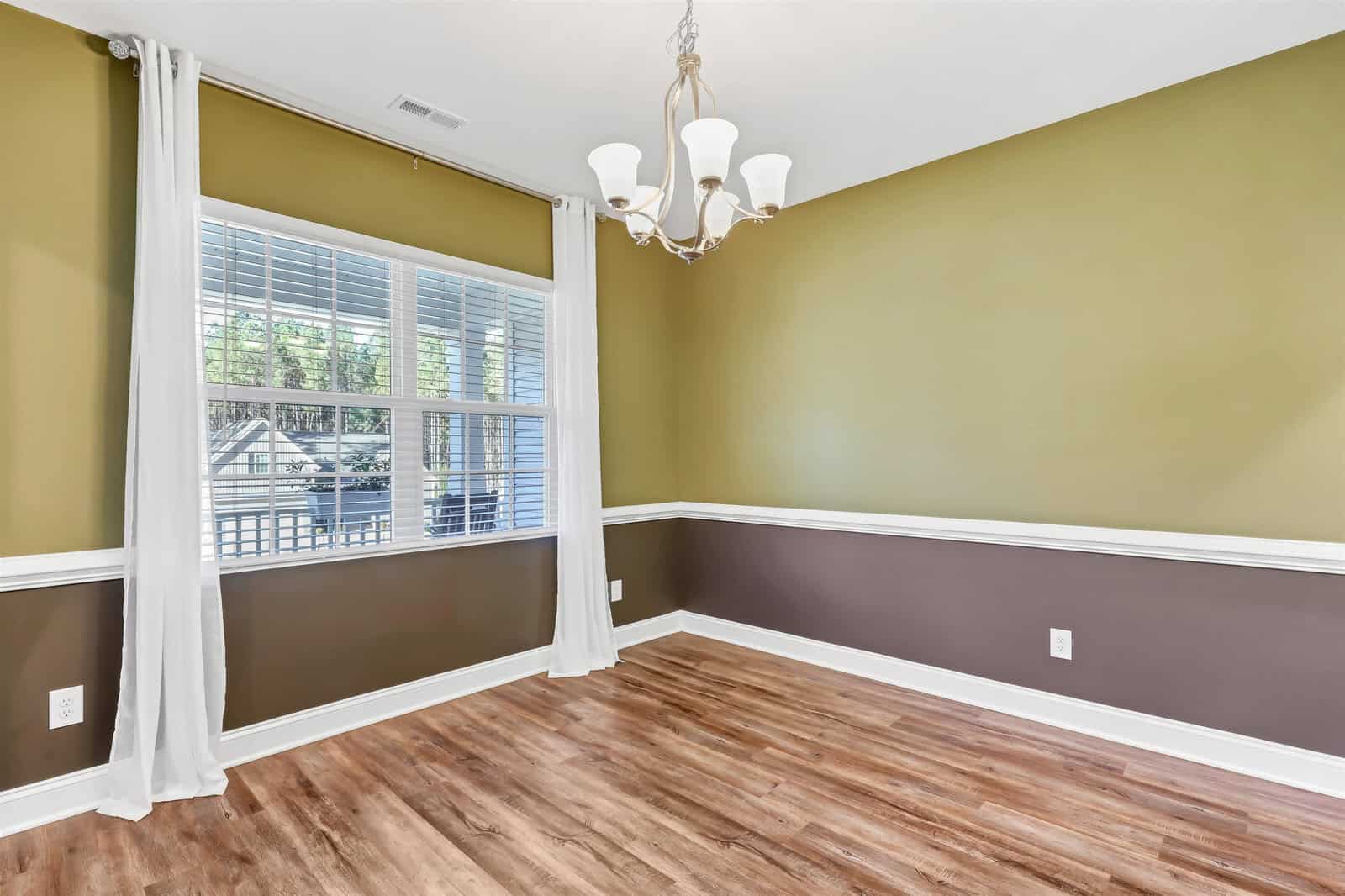 An empty room with green walls and a brown accent, featuring a large window with white curtains, a modern chandelier, and wood-patterned flooring.