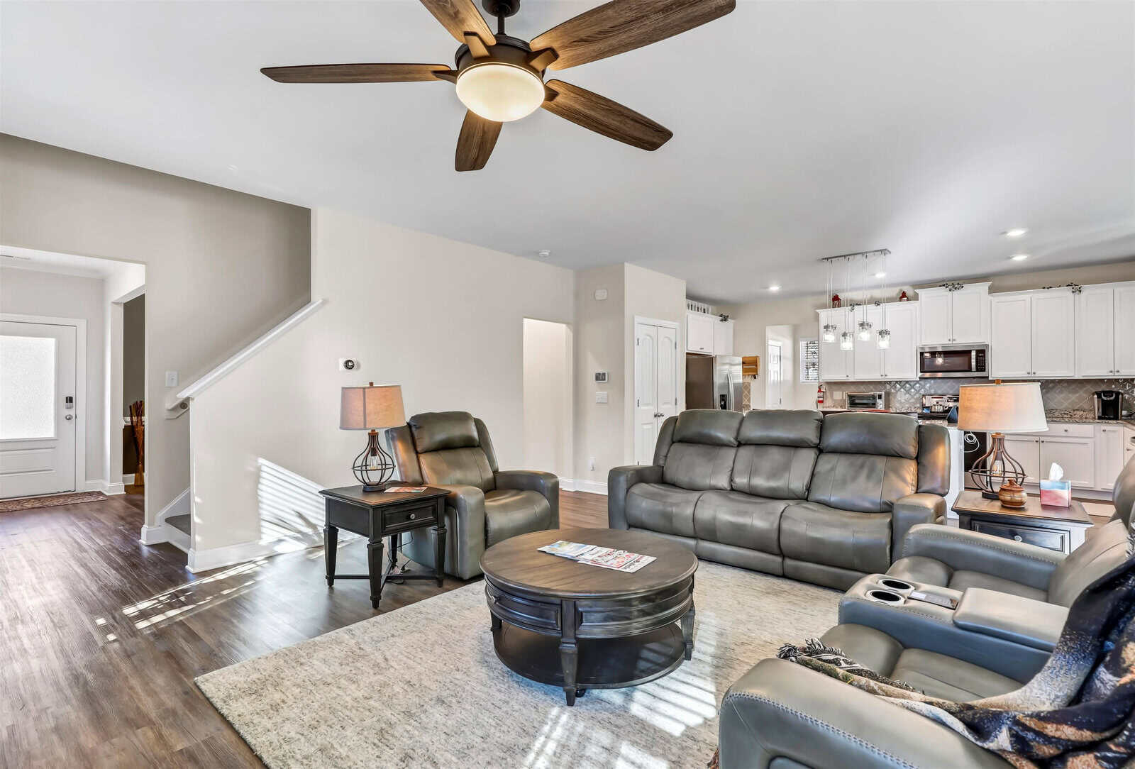 A modern living room with gray leather sofas and recliners arranged around a round wooden coffee table. A ceiling fan is above, and the room opens to a kitchen with white cabinetry. Sunlight filters through windows, highlighting the hardwood floor.
