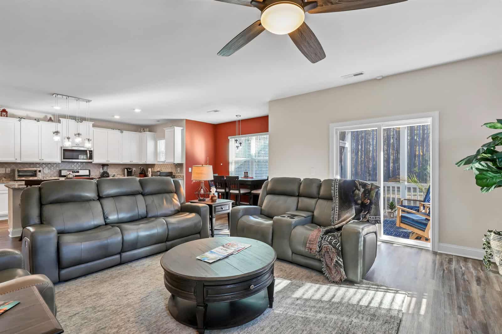 A spacious living room with dark leather sofas, a round coffee table, and an overhead ceiling fan. The room is open to a kitchen area with white cabinets. Sunlight streams through a glass door leading to a deck with chairs outside.