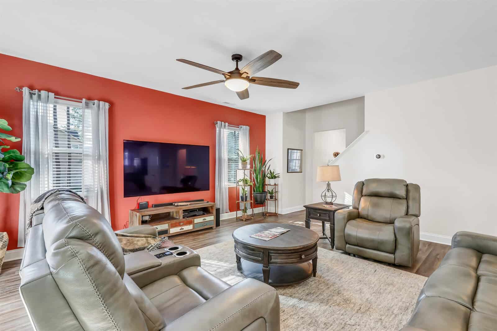 A modern living room featuring gray leather recliners, a large flat-screen TV on an orange accent wall, and a round coffee table. The room has light wood flooring, a ceiling fan, and natural light coming through two windows with sheer curtains.