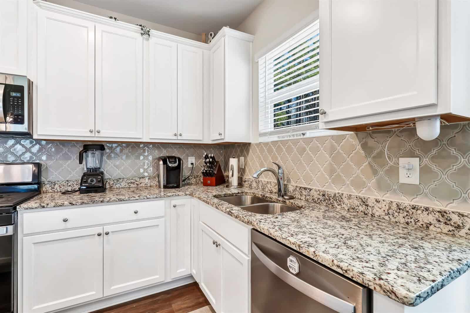 A modern kitchen with white cabinets and a tiled backsplash features a stainless steel sink, dishwasher, and granite countertops. Appliances including a blender, coffee maker, and knife set are on the countertop. A window adds natural light.