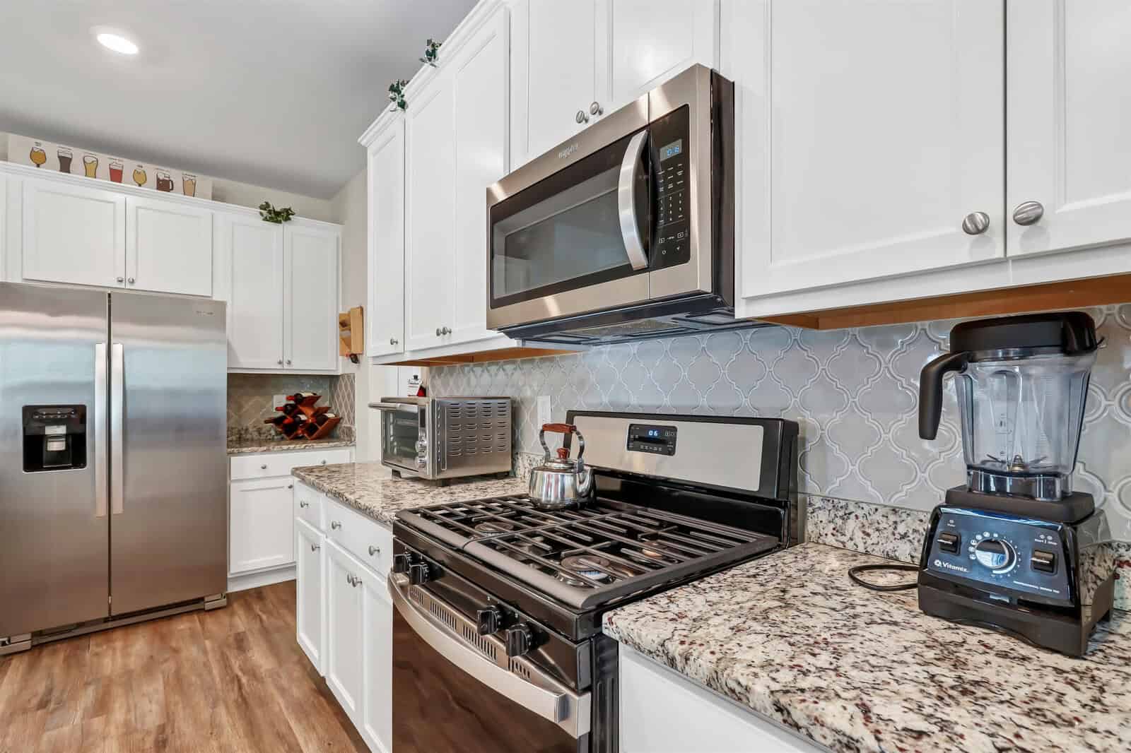 A modern kitchen featuring white cabinets, stainless steel appliances, and a granite countertop. A gas stove with a kettle, a microwave, a blender, and a toaster oven are visible. A fridge and decorative items are in the background.