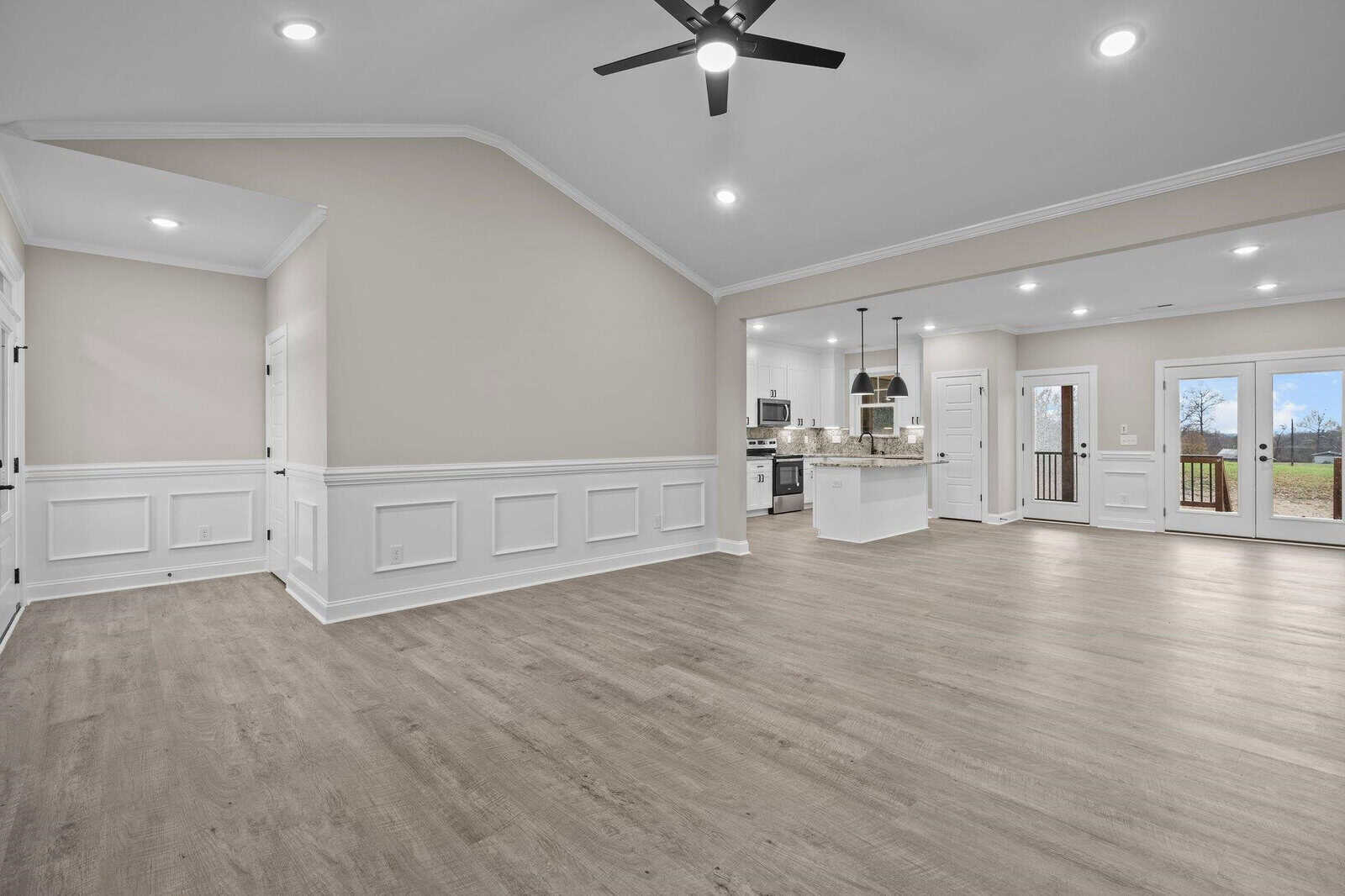 A spacious, empty living room with hardwood floors leading into an open kitchen with modern appliances and white cabinets. the room features recessed lighting, a ceiling fan, white wainscoting, and glass doors that provide natural light.