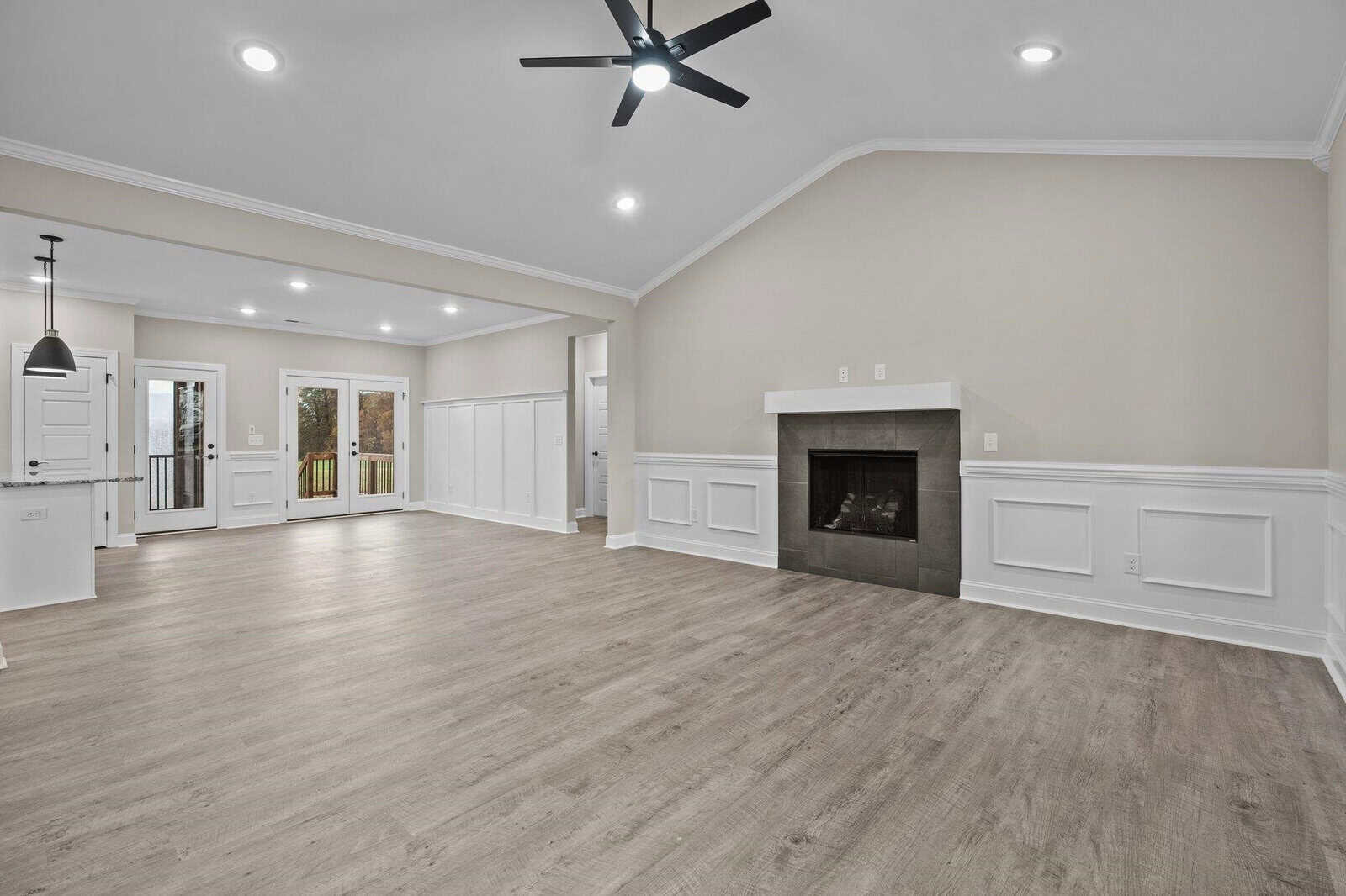 A spacious, empty room featuring a modern fireplace, hardwood floors, white wainscoting, and recessed ceiling lighting, with an open doorway leading to another room and french doors at the rear.