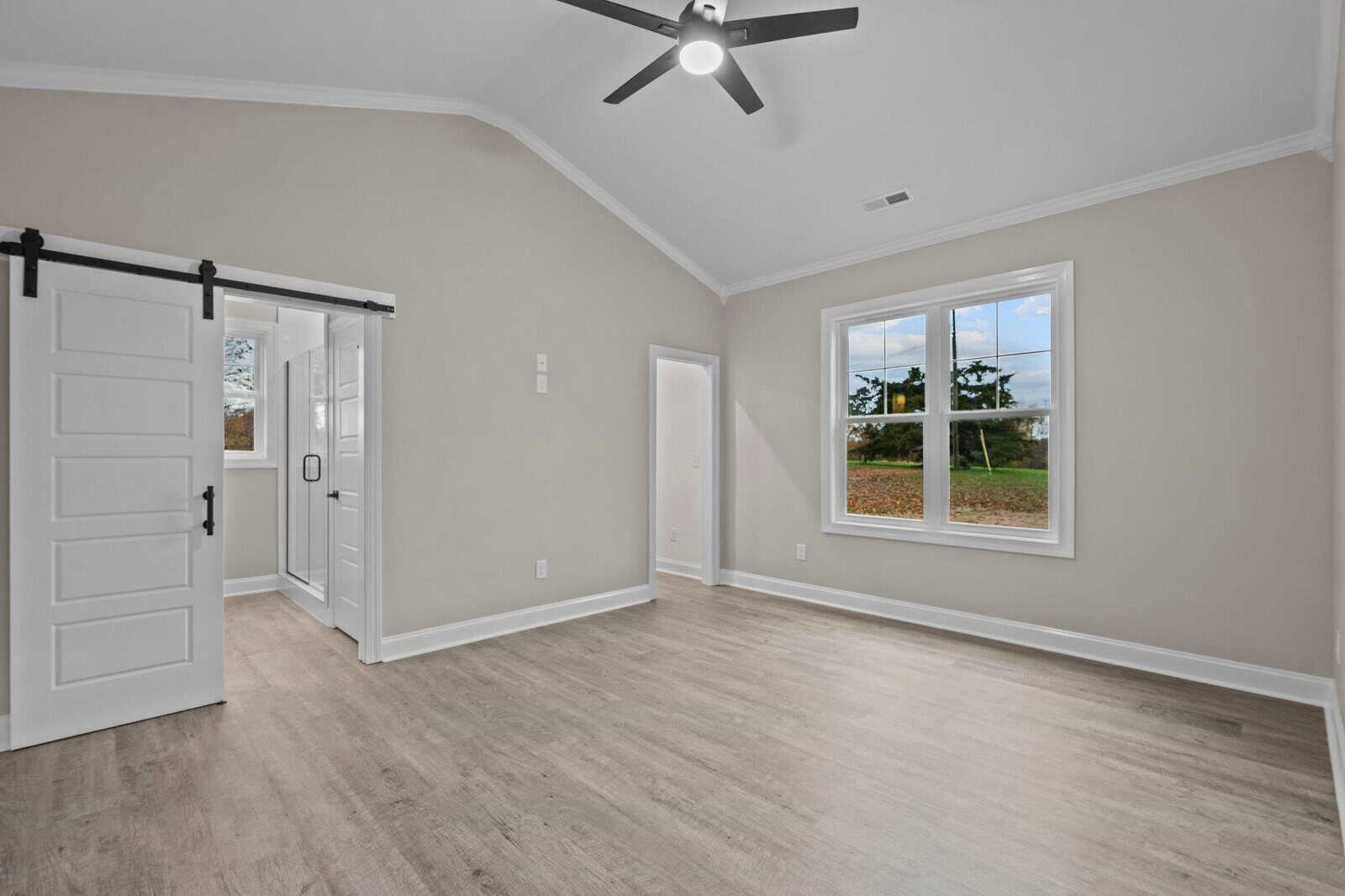 A spacious, empty room with a vaulted ceiling, featuring a black ceiling fan, modern sliding barn door, and a large window allowing natural light to enter.