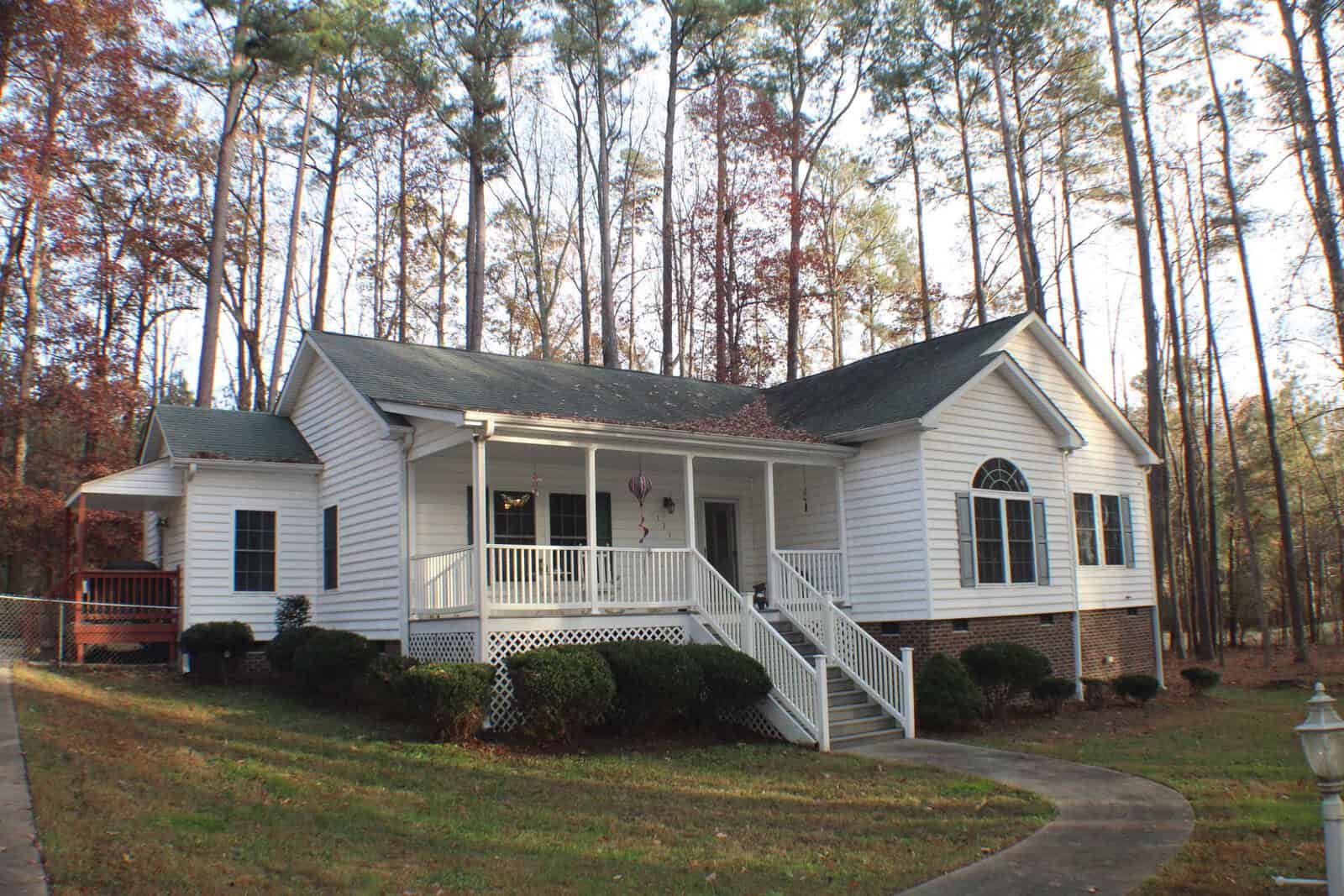 A white single-story house at 135 S S Chaucer Way, featuring a front porch and steps leading to the entrance, is situated amidst tall trees. The Kittrell residence has a gable roof and is surrounded by a well-kept lawn and bushes. The setting appears peaceful with woodland scenery in the background.