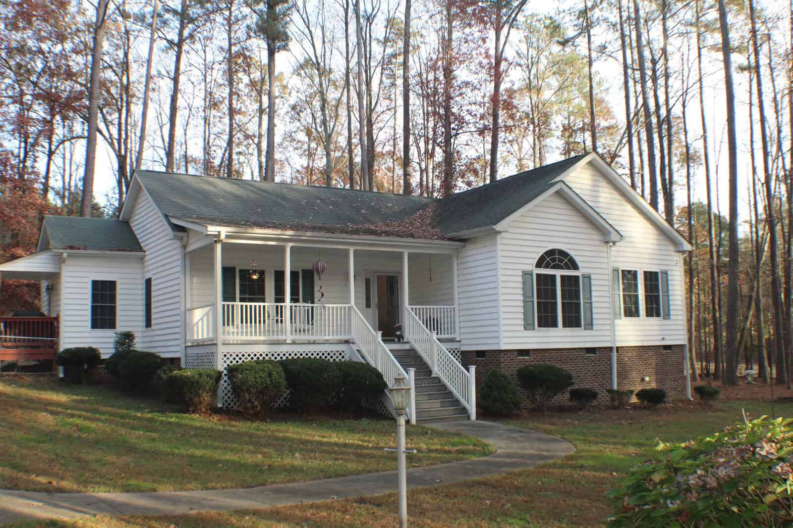 A single-story white house with a spacious front porch and a brick foundation, surrounded by tall trees and a lawn. The home features several large windows and a set of steps leading up to the front door. A curved walkway leads from the front door to the yard.
