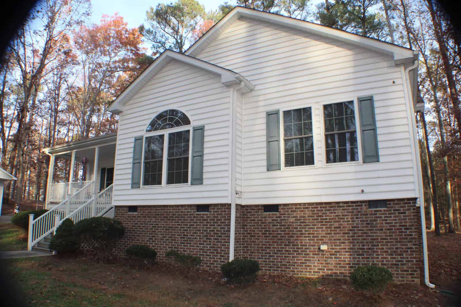 A white house with light green shutters features a combination of brick and siding exterior. The house has a large, arched window on the front and a small set of steps leading to a covered porch. It is surrounded by a grassy yard and tall trees.