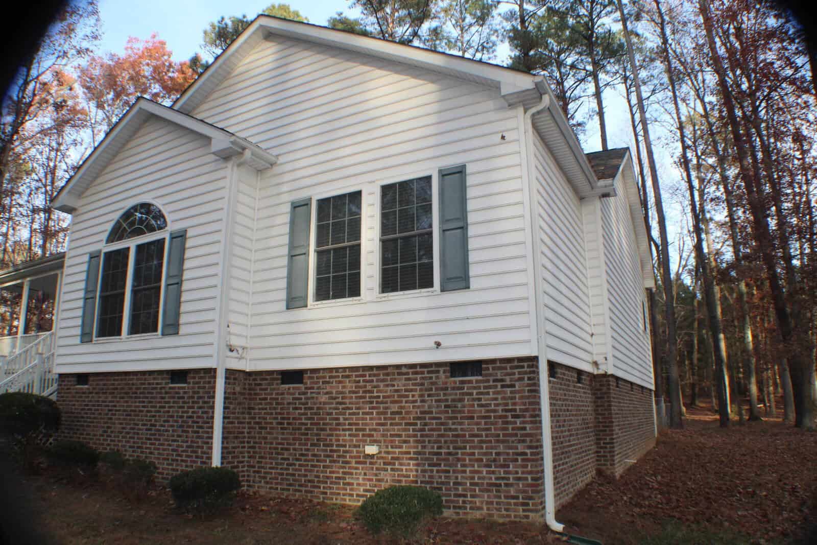 A two-story house with white siding and a brick foundation is surrounded by tall trees. The side of the house features large windows with shutters, and a downspout is visible on the right corner. The yard has trimmed shrubs and fallen leaves on the ground.