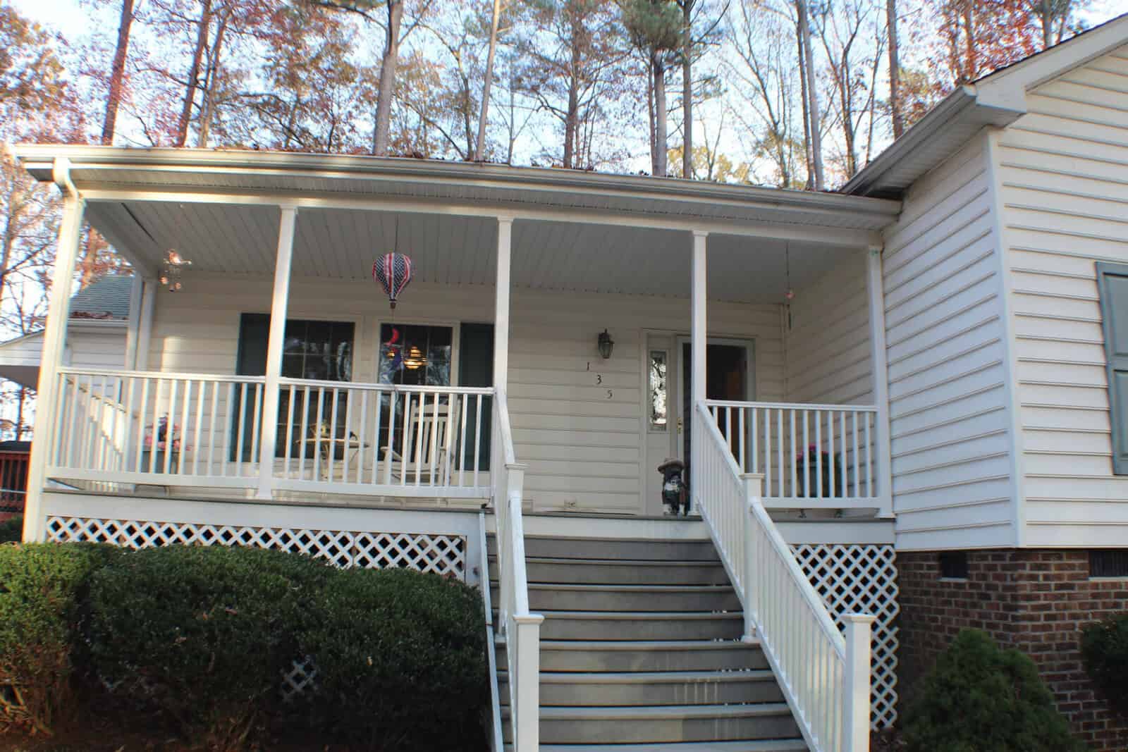 A white, two-story house with an open front porch and a staircase leading up to it. The porch is adorned with hanging decorations and potted plants. The house is surrounded by trees with autumn leaves.