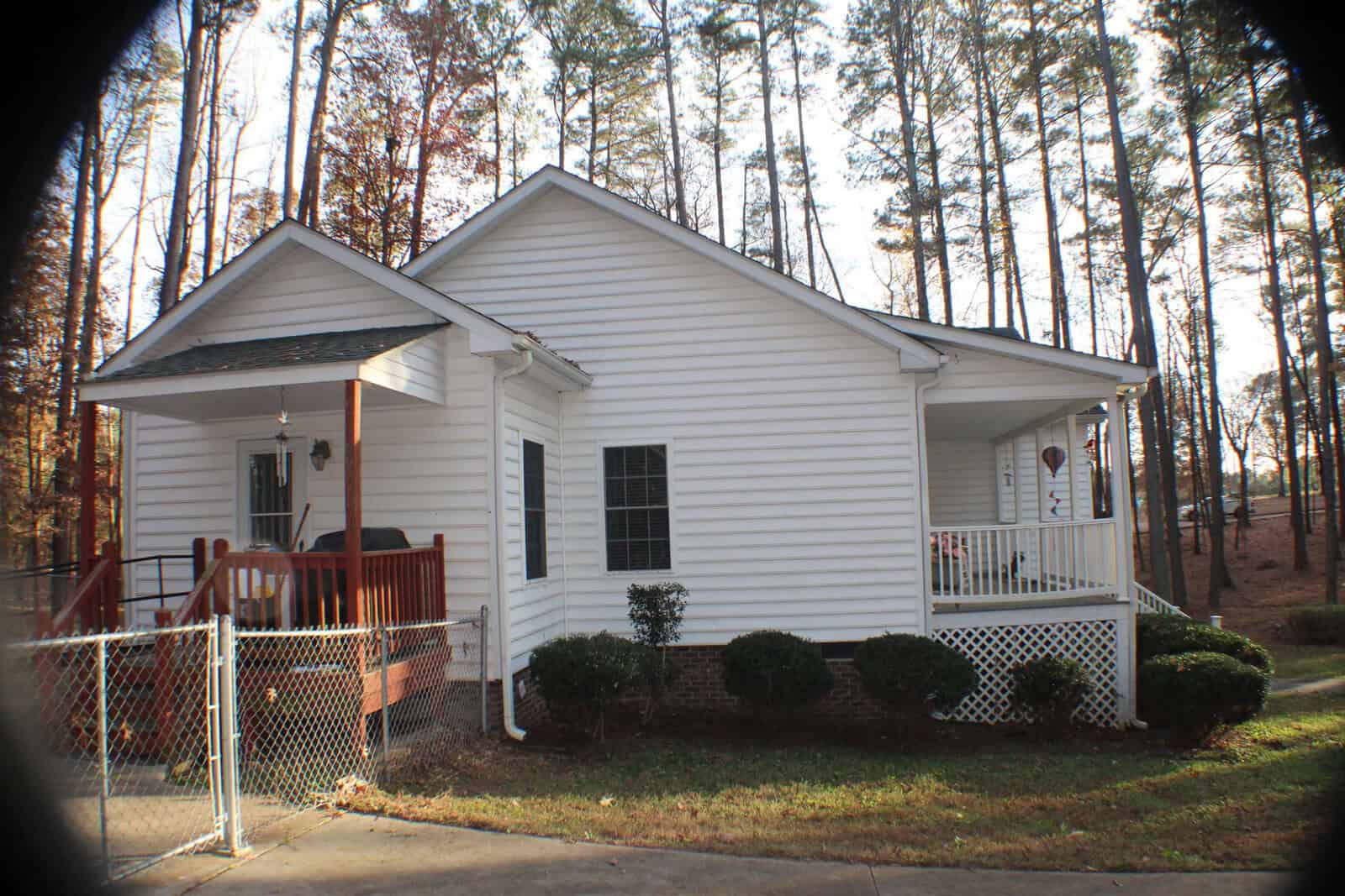 A white single-story house with a green roof is surrounded by tall trees. The home has a small front porch with railings, a covered side patio, and a chain-link fence to the left. Shrubs are neatly planted along the foundation. The setting appears peaceful.